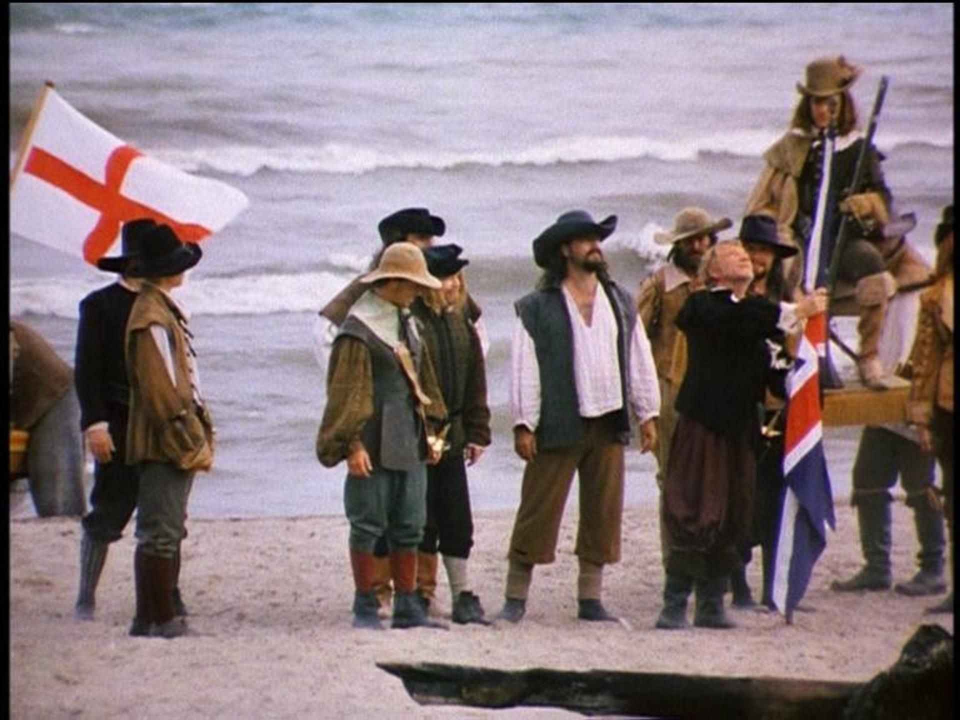 Männer in historischer Kleidung am Strand mit Flaggen. Ein Mann hält eine Flagge mit rotem Kreuz. Wellen im Hintergrund.