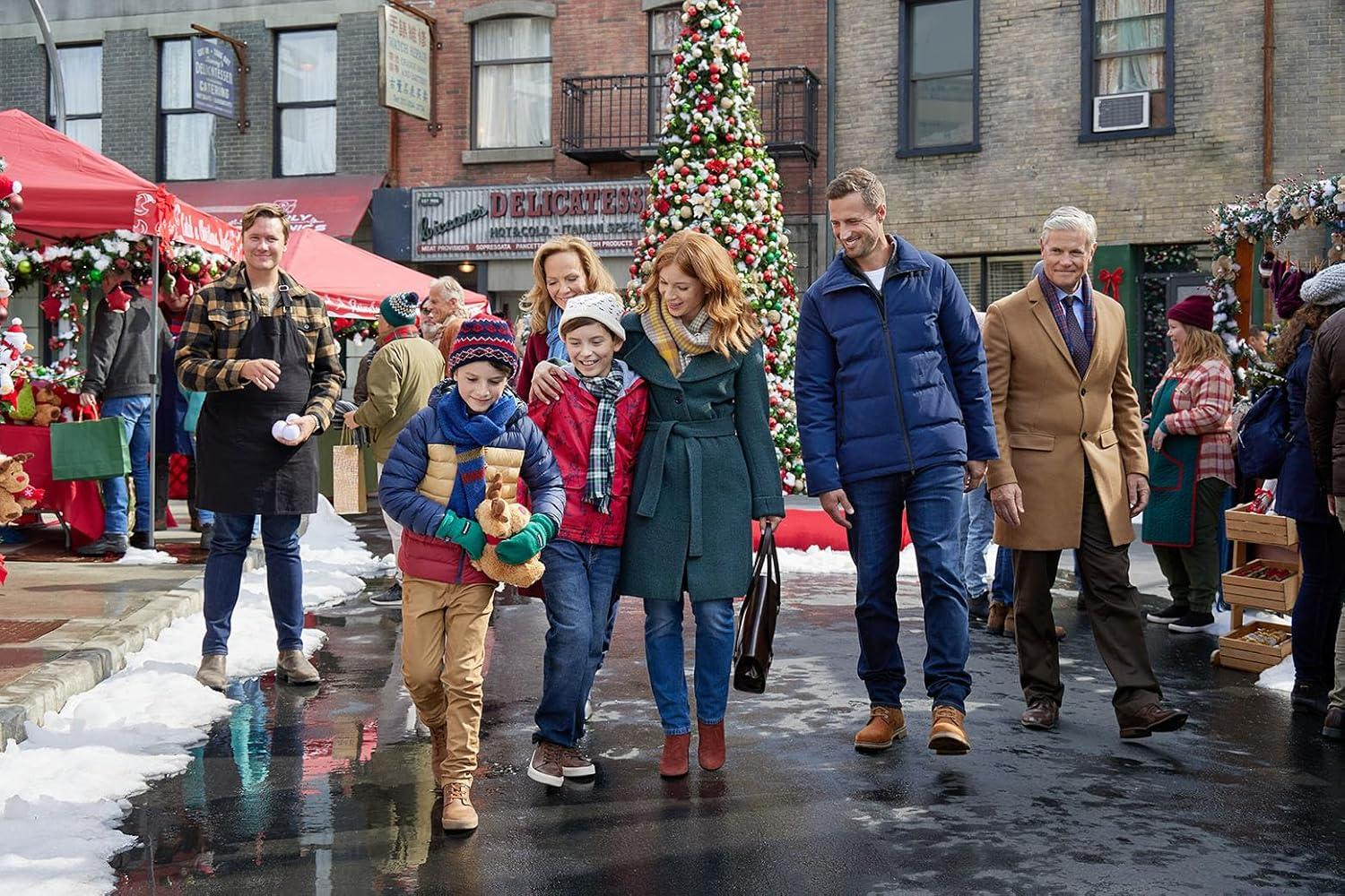 Eine Familie geht vor einem Weihnachtsmarkt auf einer nassen Straße. Hinter ihnen steht ein Baum. Leute sind beim Einkaufen.