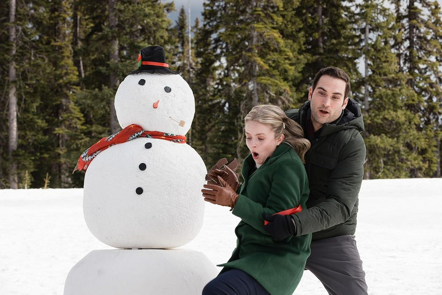 Ein Schneemann mit Hut und Schal steht im Schnee. Ein Mann und eine Frau reagieren in der Nähe des Schneemanns, mit einem Wald im Hintergrund.