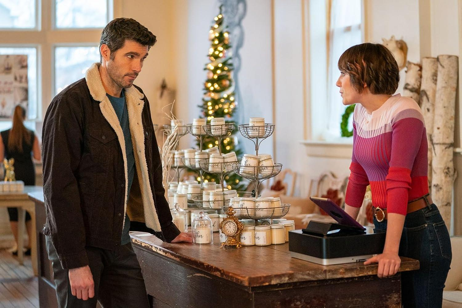 Ein Mann und eine Frau stehen an einer Theke in einem Geschäft. Kerzen werden ausgestellt. Im Hintergrund ist ein Weihnachtsbaum zu sehen.