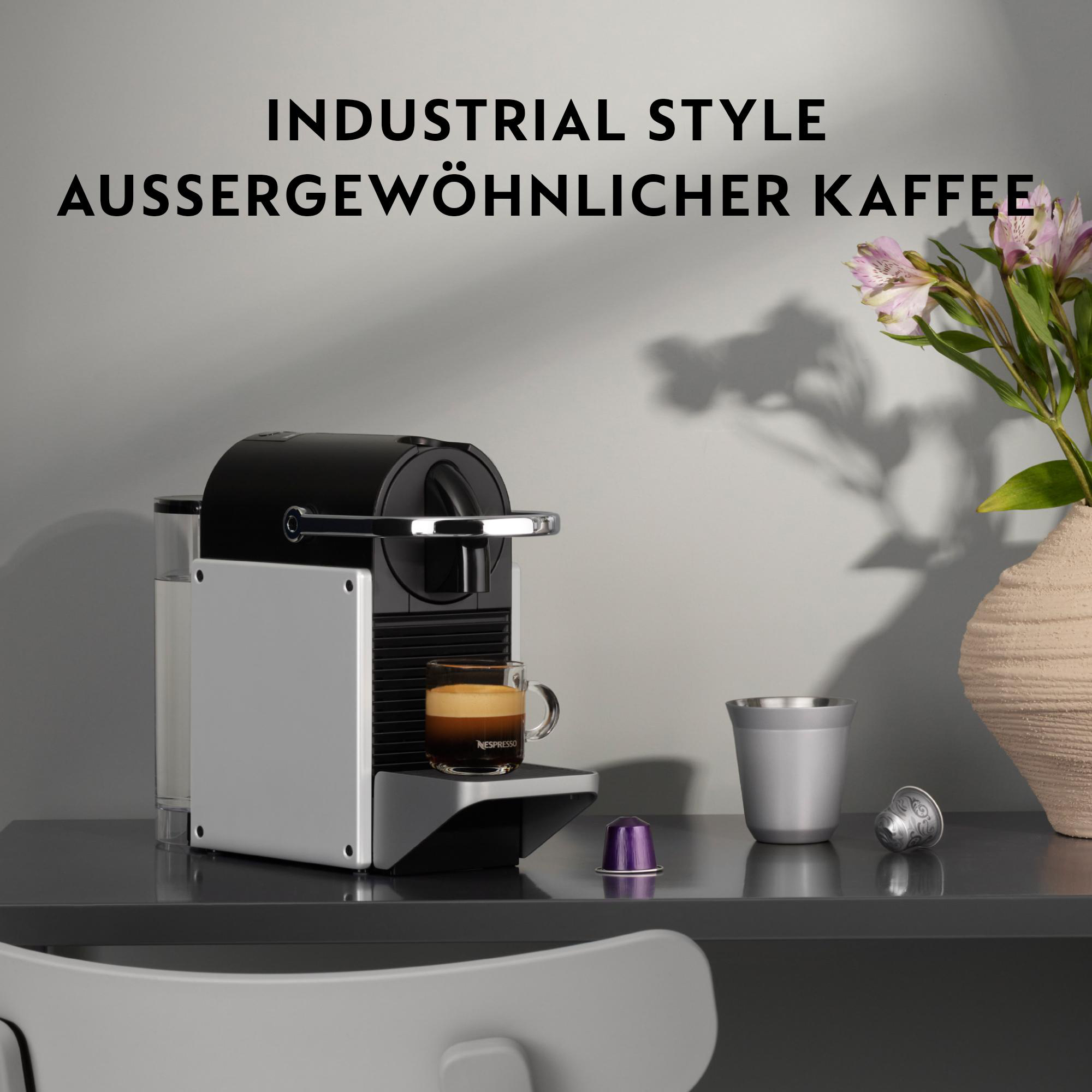 Eine Kaffeemaschine im Industriestil mit einem Glas Espresso, Blumen in einer Vase und einer Tasse. Die Wörter sind auf Deutsch.