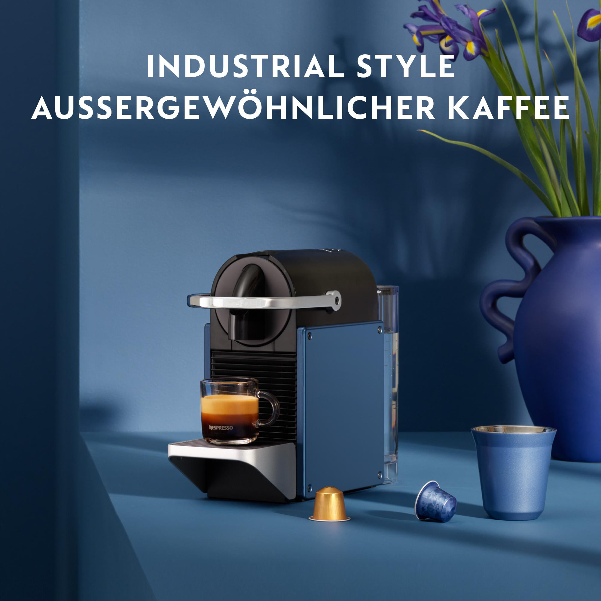 Kaffeemaschine mit einem Glas Kaffee, Kapseln und einer blauen Tasse auf einer blauen Oberfläche. Vase mit Blumen im Hintergrund.