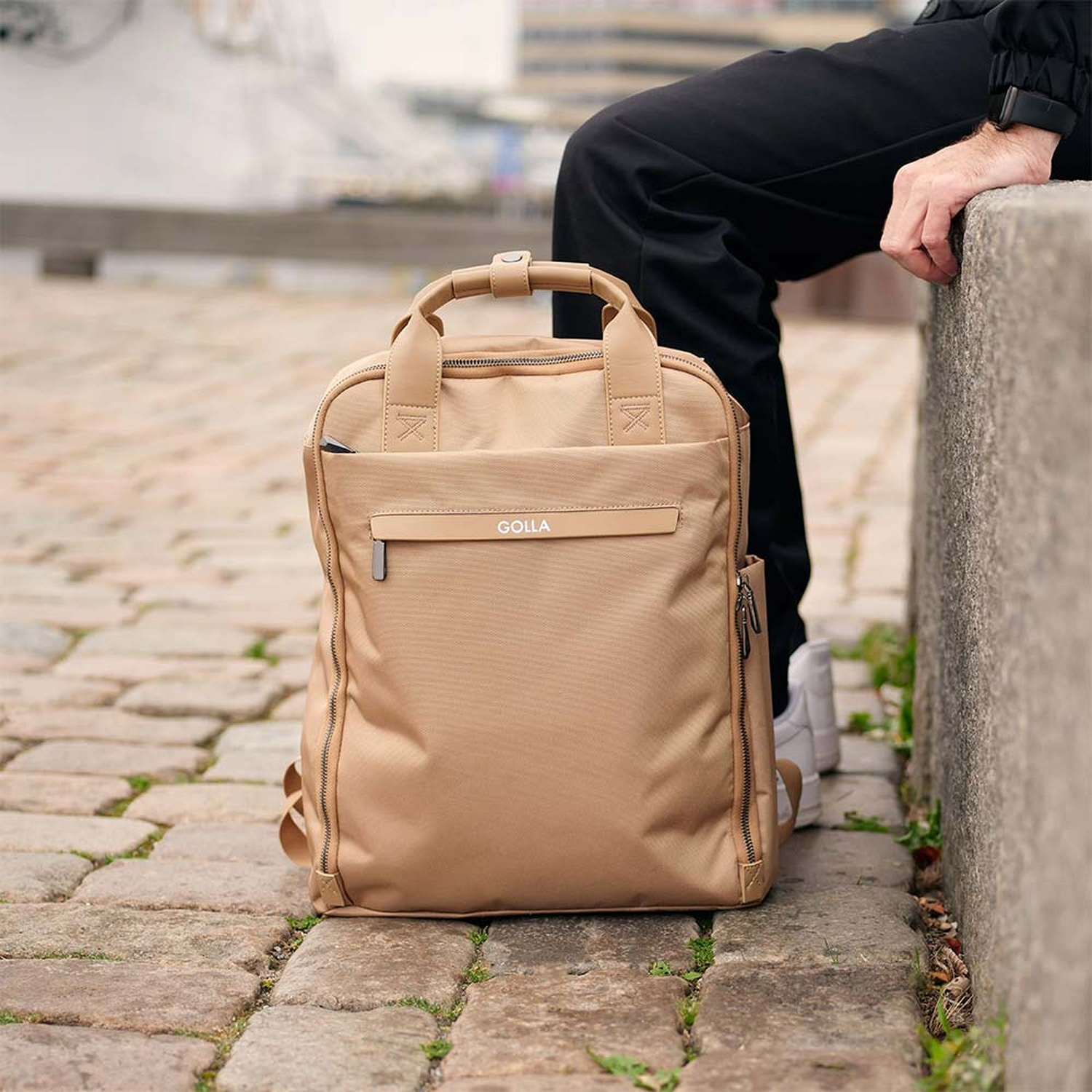 Uno zaino color cachi su ciottoli. Una persona con pantaloni neri e scarpe bianche è accanto alla borsa. La persona è appoggiata a un muro di cemento.
