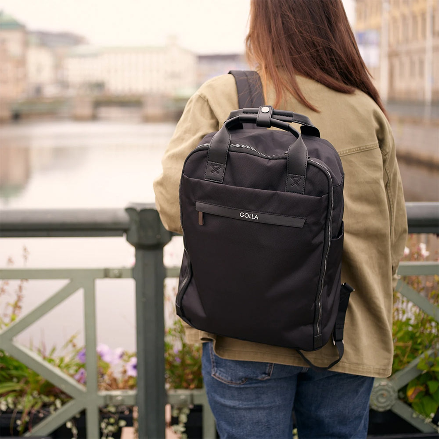 Una persona è in piedi su un ponte, con uno zaino nero. Edifici e acqua sullo sfondo. Lo zaino è della marca GOLLA.