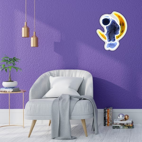 Naklejka z astronautą jest na fioletowej ścianie. Szary fotel i stolik z rośliną są z przodu. Z sufitu zwisają złote lampy.
