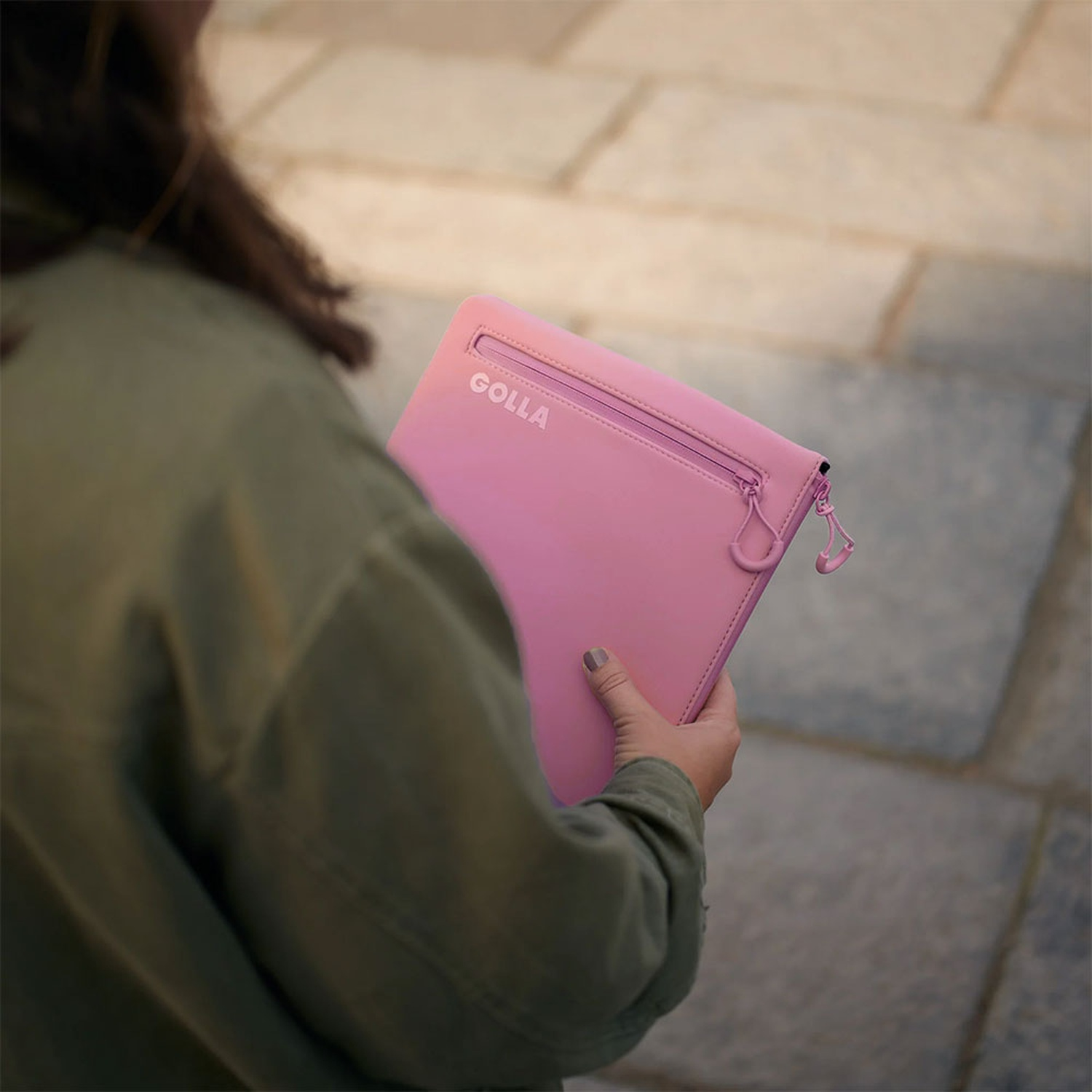 Una persona tiene in mano una borsa GOLLA rosa con cerniera. Sono all'aperto su una superficie pavimentata. La persona indossa una giacca verde.