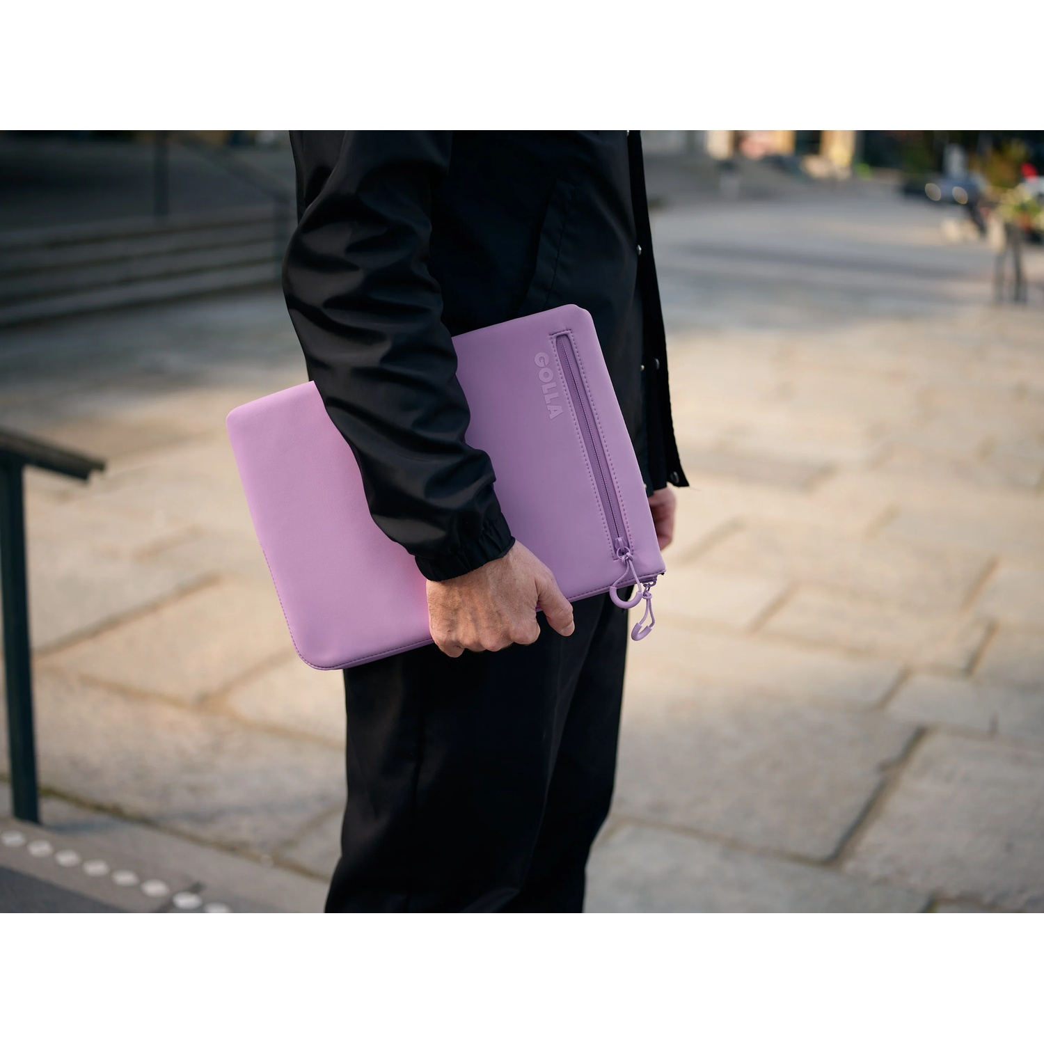 Una persona tiene in mano una custodia per laptop lilla. La persona indossa abiti neri. Lo sfondo è una scena sfocata all'aperto con pietre.