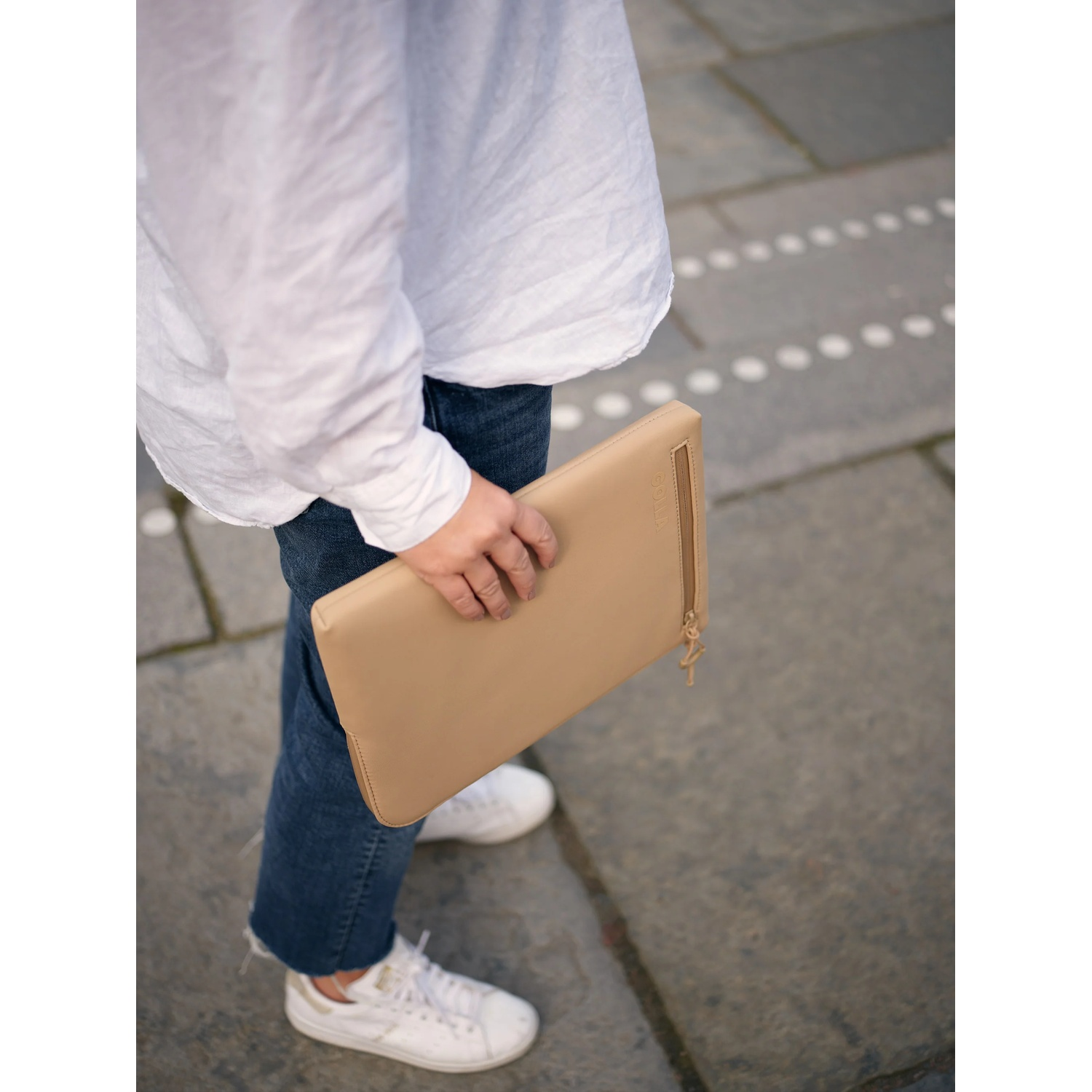 Una persona che tiene una pochette beige, indossa una camicia bianca, jeans e scarpe da ginnastica bianche, in piedi su una superficie di pietra.