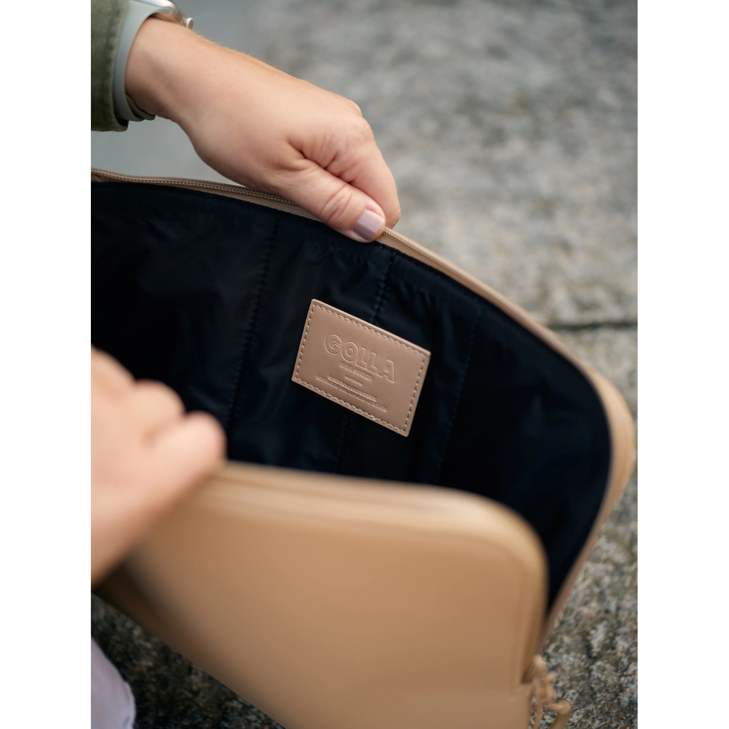 Le mani aprono una borsa beige con interno nero. Un'etichetta dice 'GOLLA' al centro. Lo sfondo è sfocato.