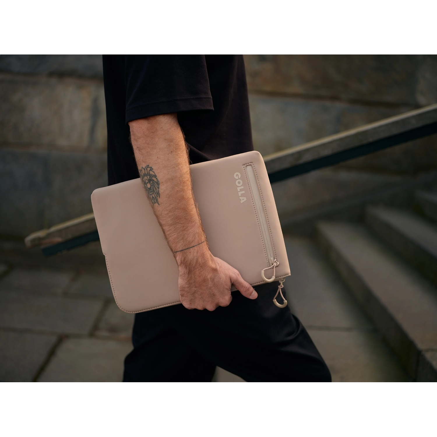 Una persona tiene in mano una custodia beige per laptop, che scende le scale. La custodia ha una cerniera e il marchio GOLLA. La persona indossa il nero.