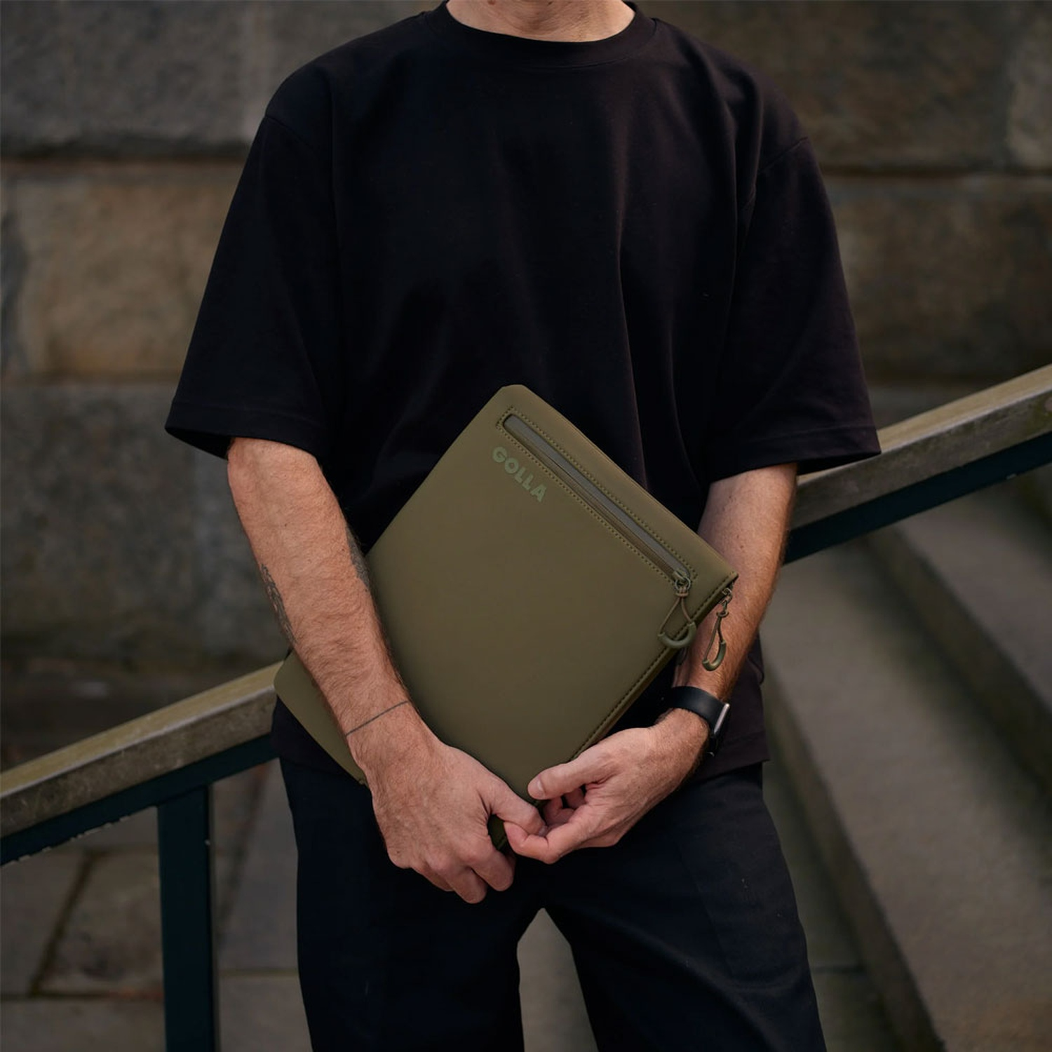 Un uomo tiene in mano una custodia verde per laptop. Indossa una maglietta e pantaloni neri. Lo sfondo mostra un muro e gradini di cemento.