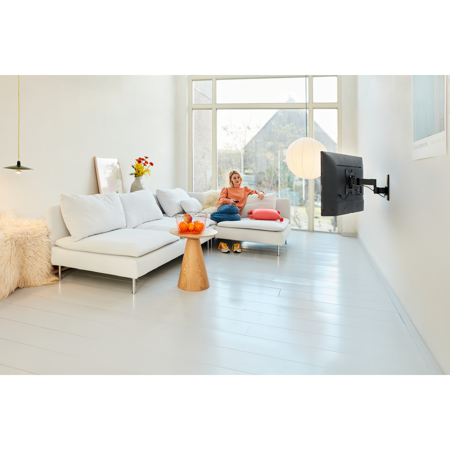 Vrouw zit op een witte bank, tv te kijken. Een tv is aan de muur bevestigd. Sinaasappels op een houten tafel. Lichte kamer.