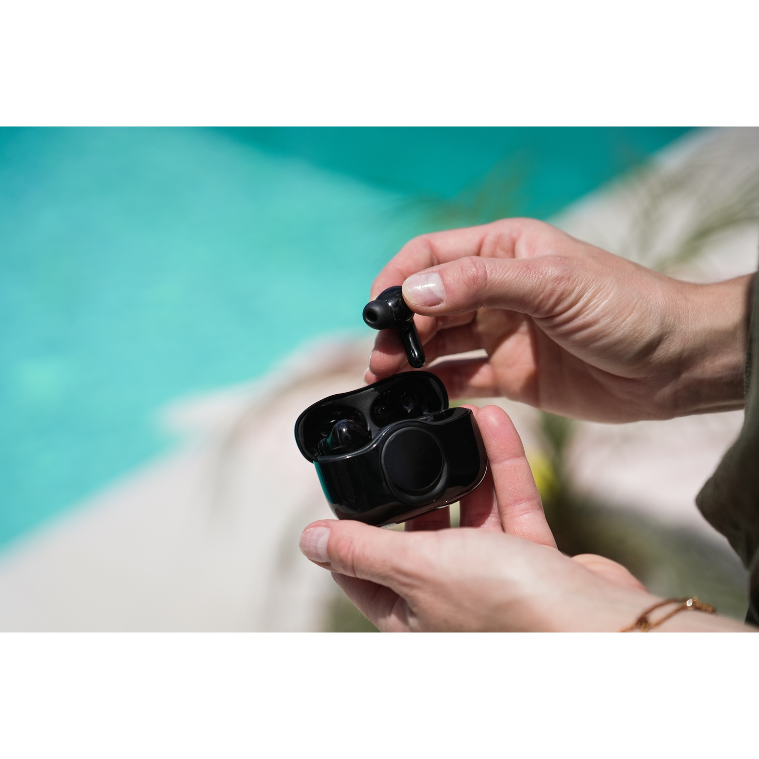 Hands holding black wireless earbuds and case. Background shows a pool with turquoise water and sunlight.