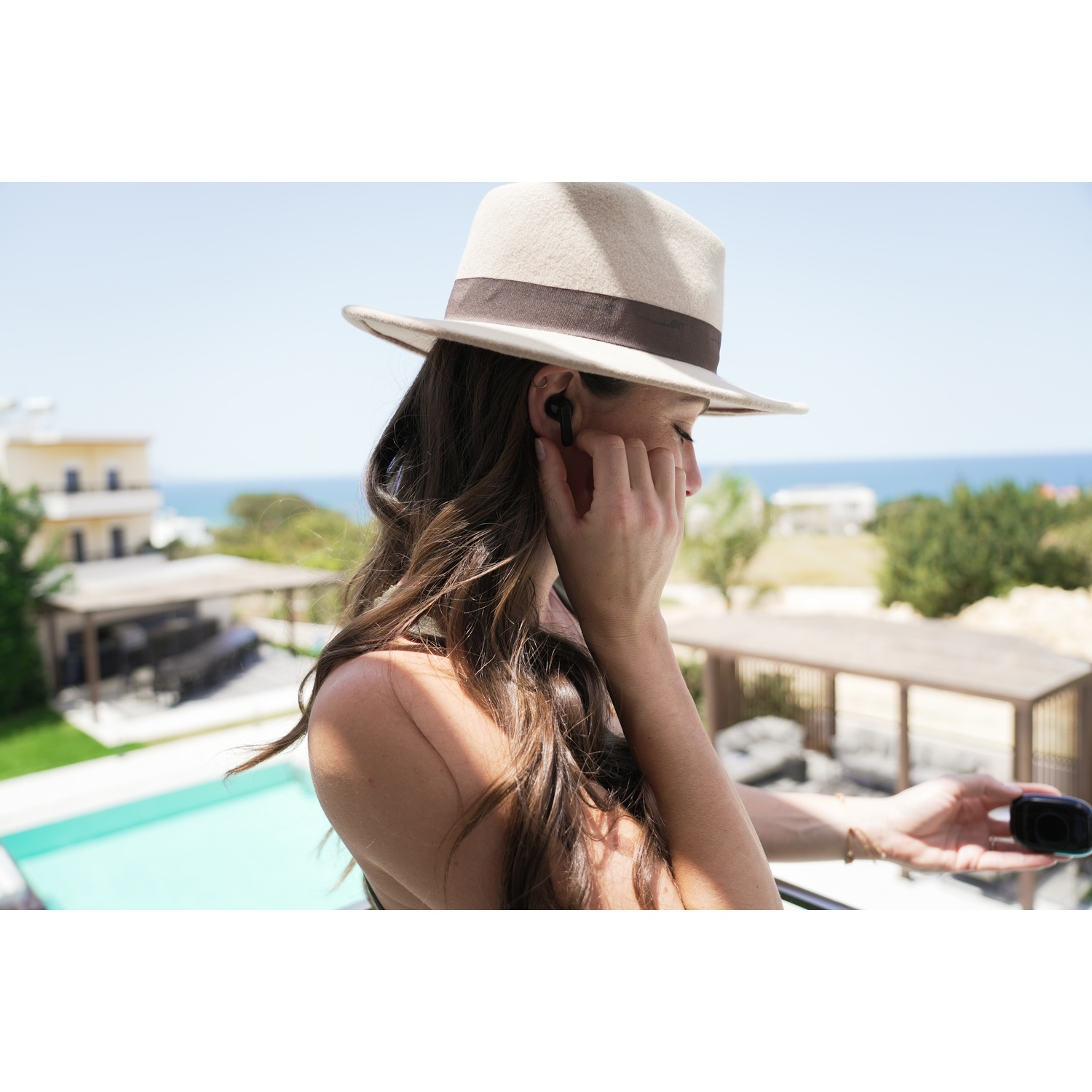 Woman wearing a hat is putting in an earbud. She is on a balcony overlooking a pool and the ocean.