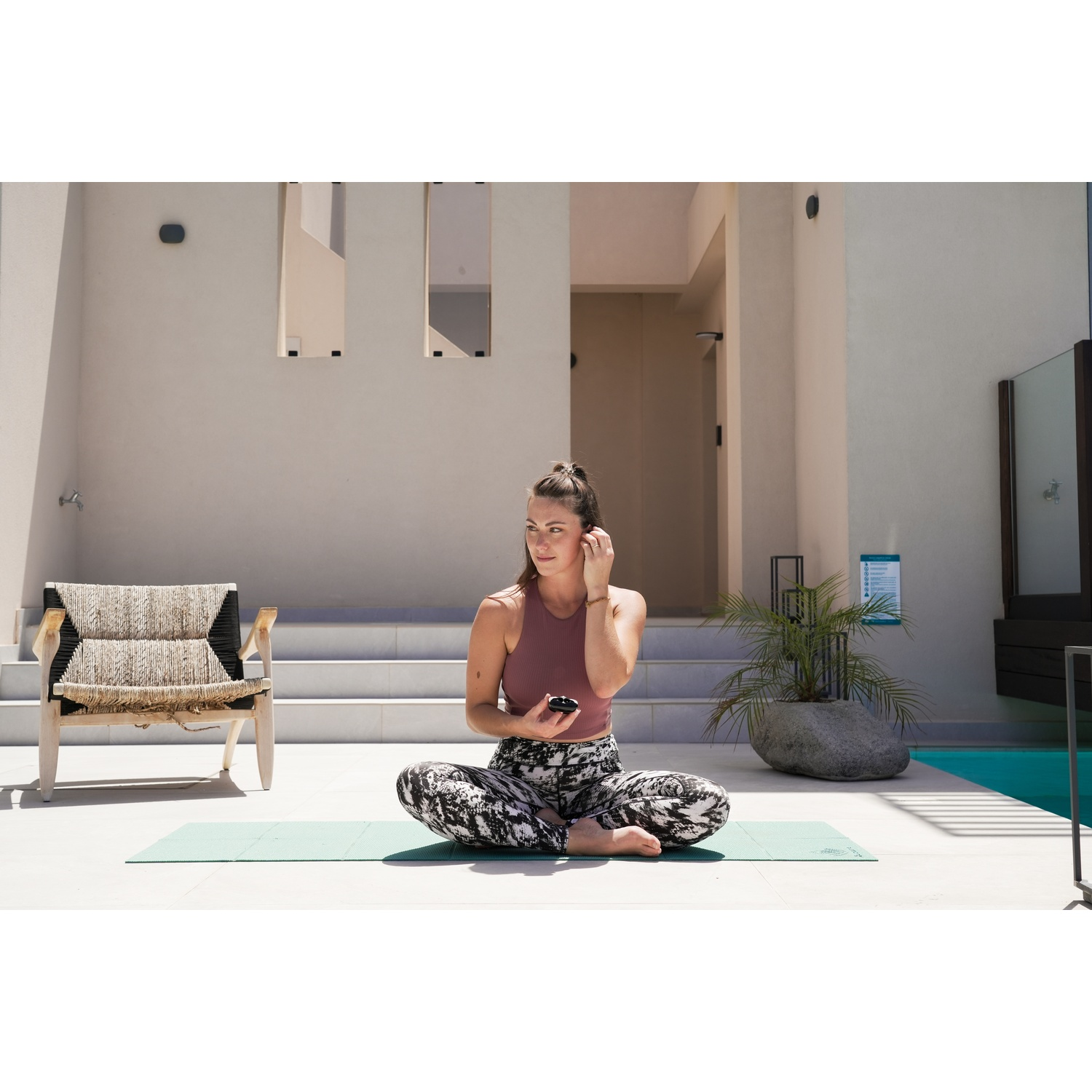 Woman in leggings and top sits on a yoga mat, holding earbud case. Background includes a pool, chair, and building.