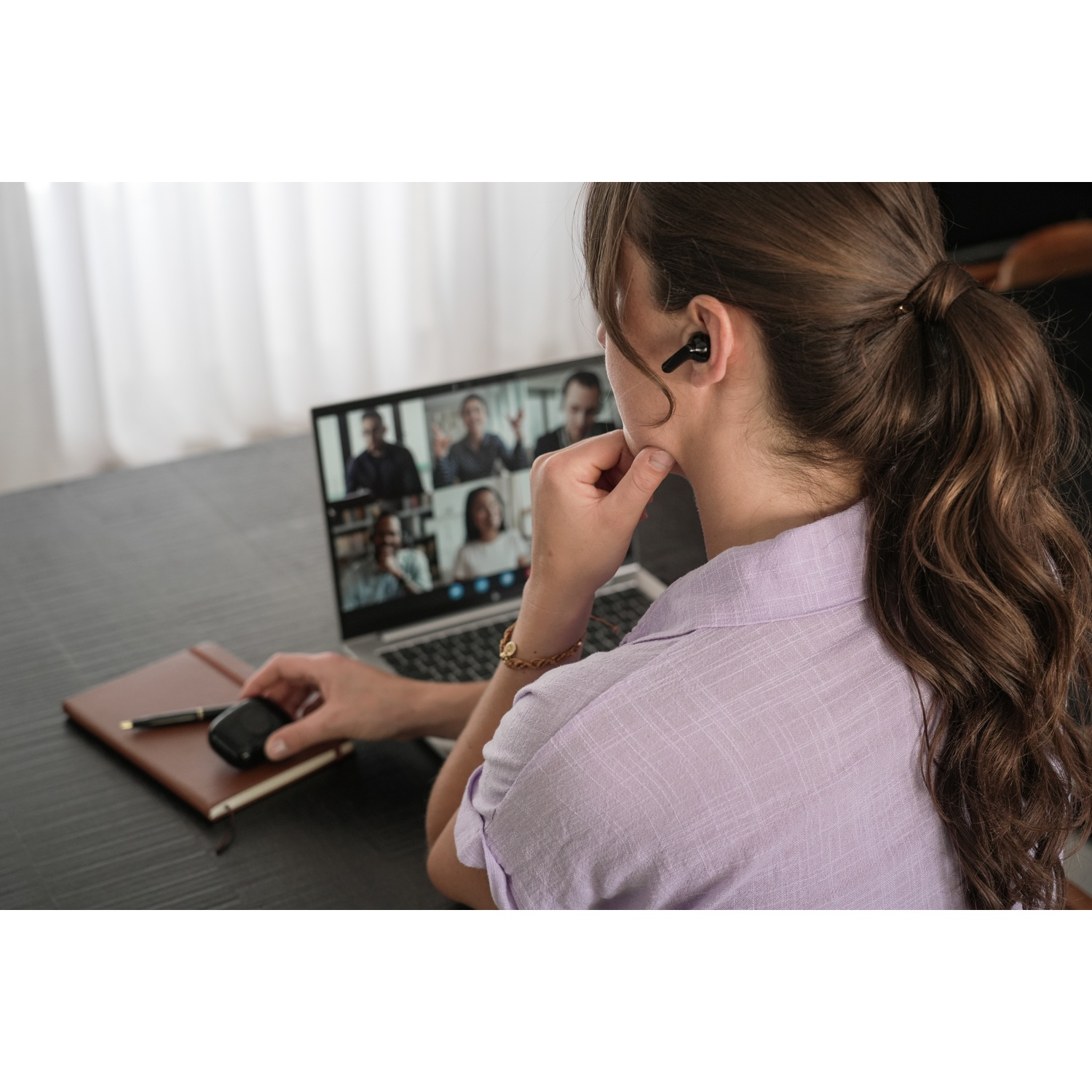 A woman with an earbud is in a video call on a laptop. She touches her face. A notebook and a mouse are on the table.