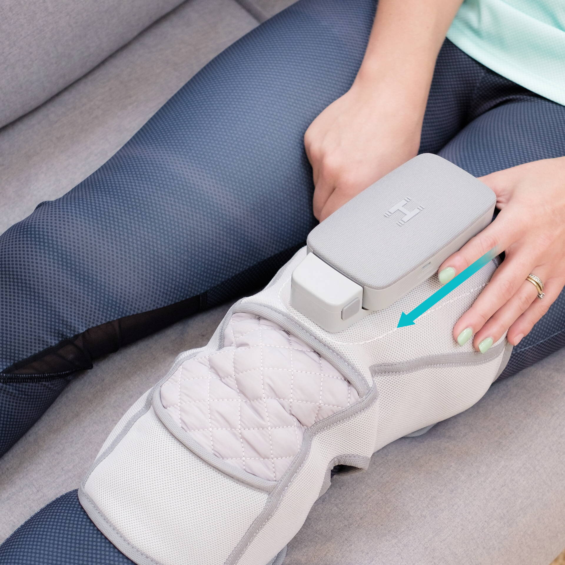 Osoba trzyma szare urządzenie na ortezi nogi. Osoba siedzi na szarej kanapie. Niebieska strzałka wskazuje w dół.