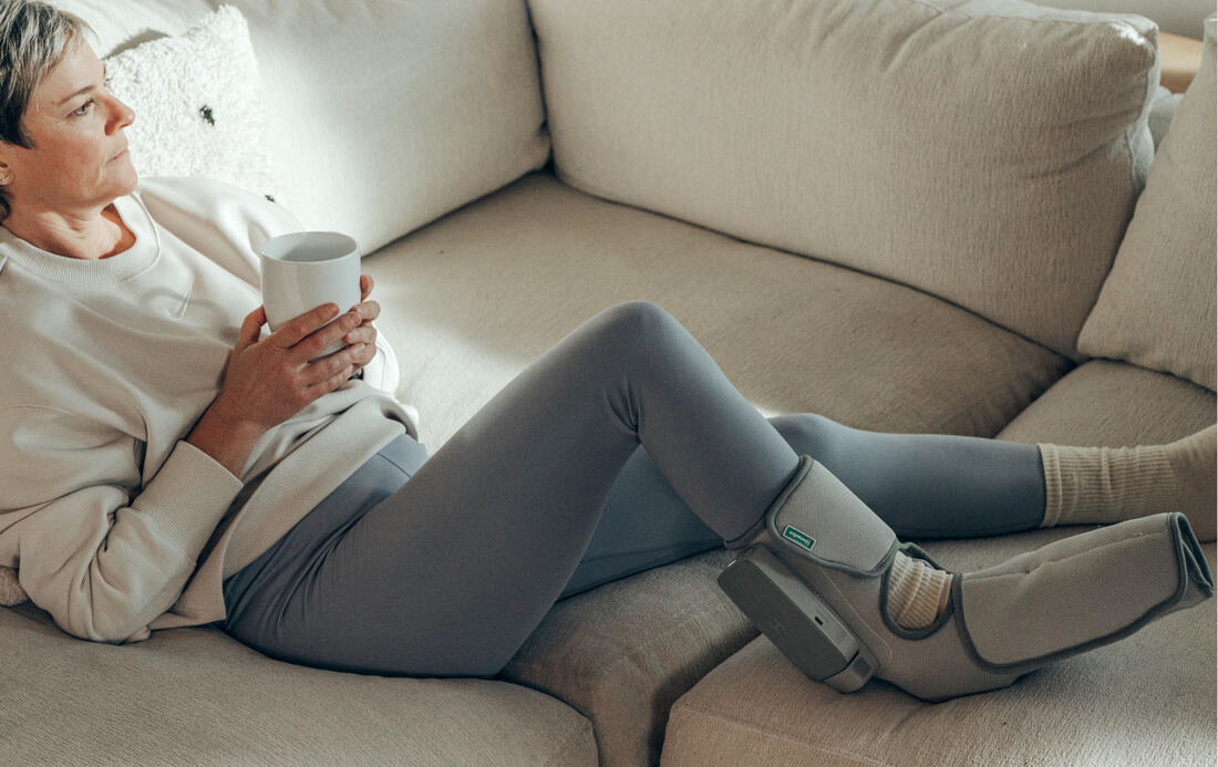 Kobieta w legginsach i swetrze relaksuje się na kanapie, trzymając kubek. Urządzenie do terapii stóp jest na jej nodze.