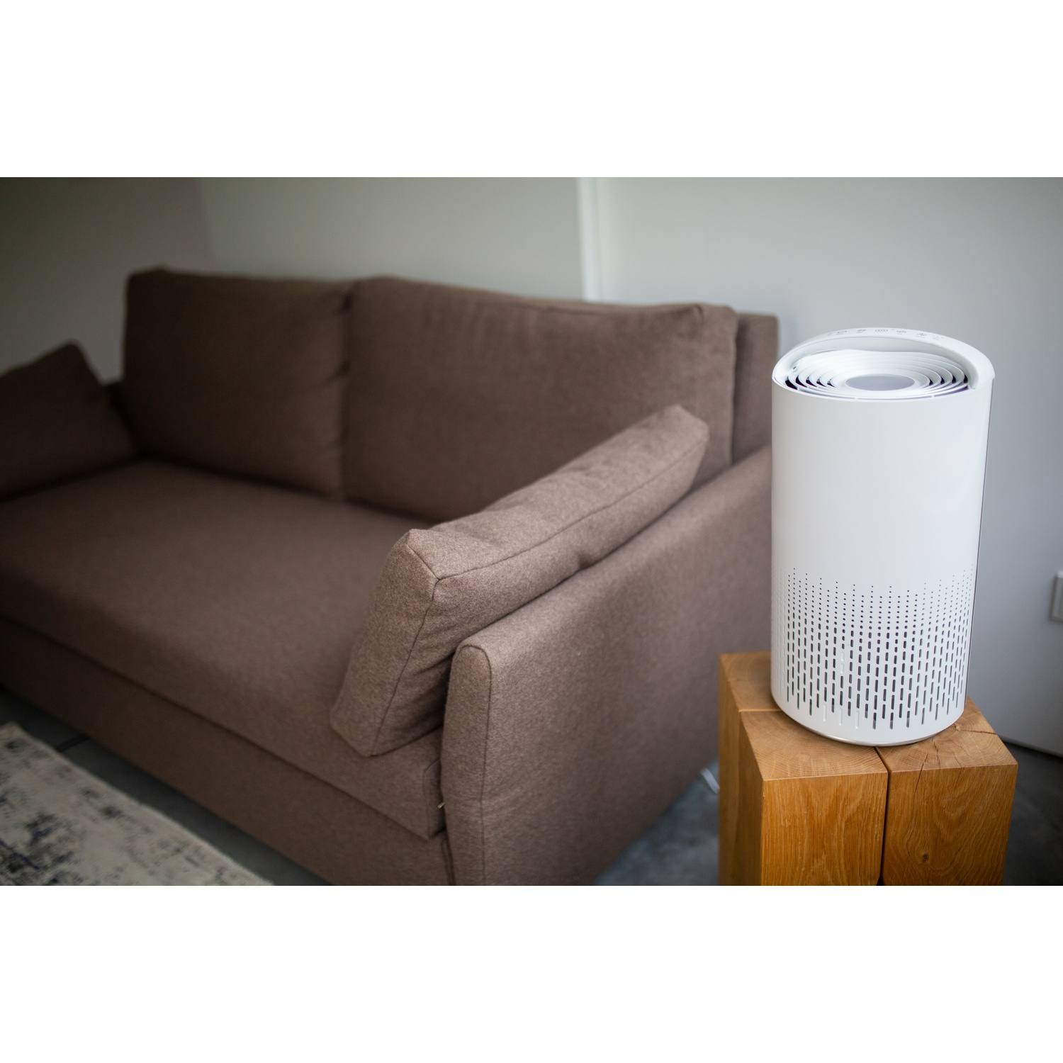 A white air purifier sits on a wooden block next to a brown sofa in a room with white walls. A rug is partially visible.