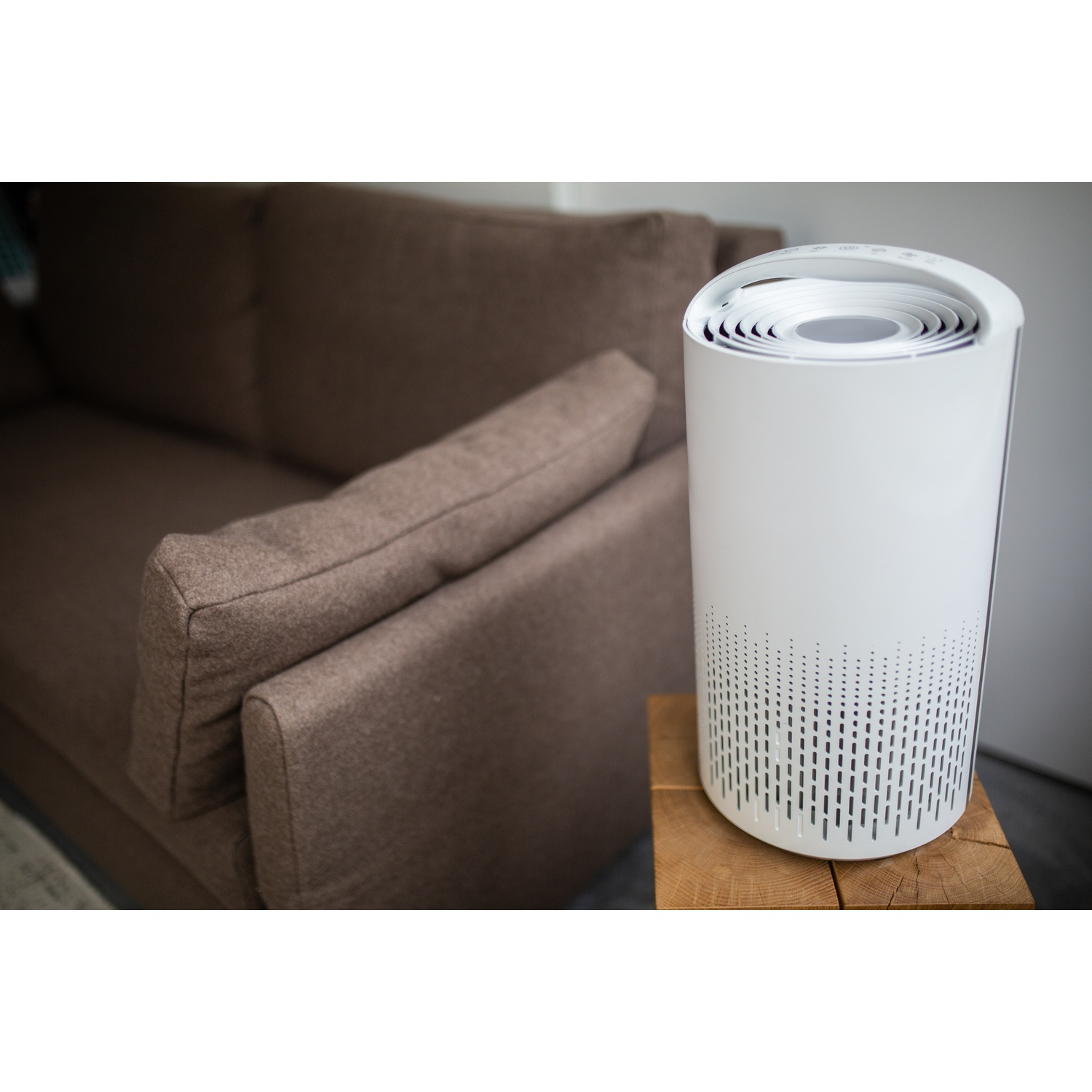 A white air purifier on a wooden table, brown sofa in the background. The purifier has a cylindrical shape.