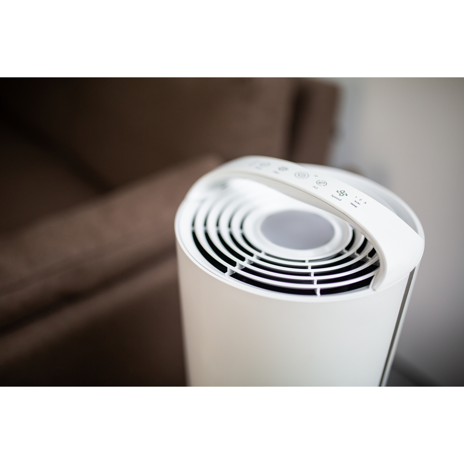 A white air purifier sits on a table. A brown sofa is in the blurred background. The purifier has a handle and control panel.