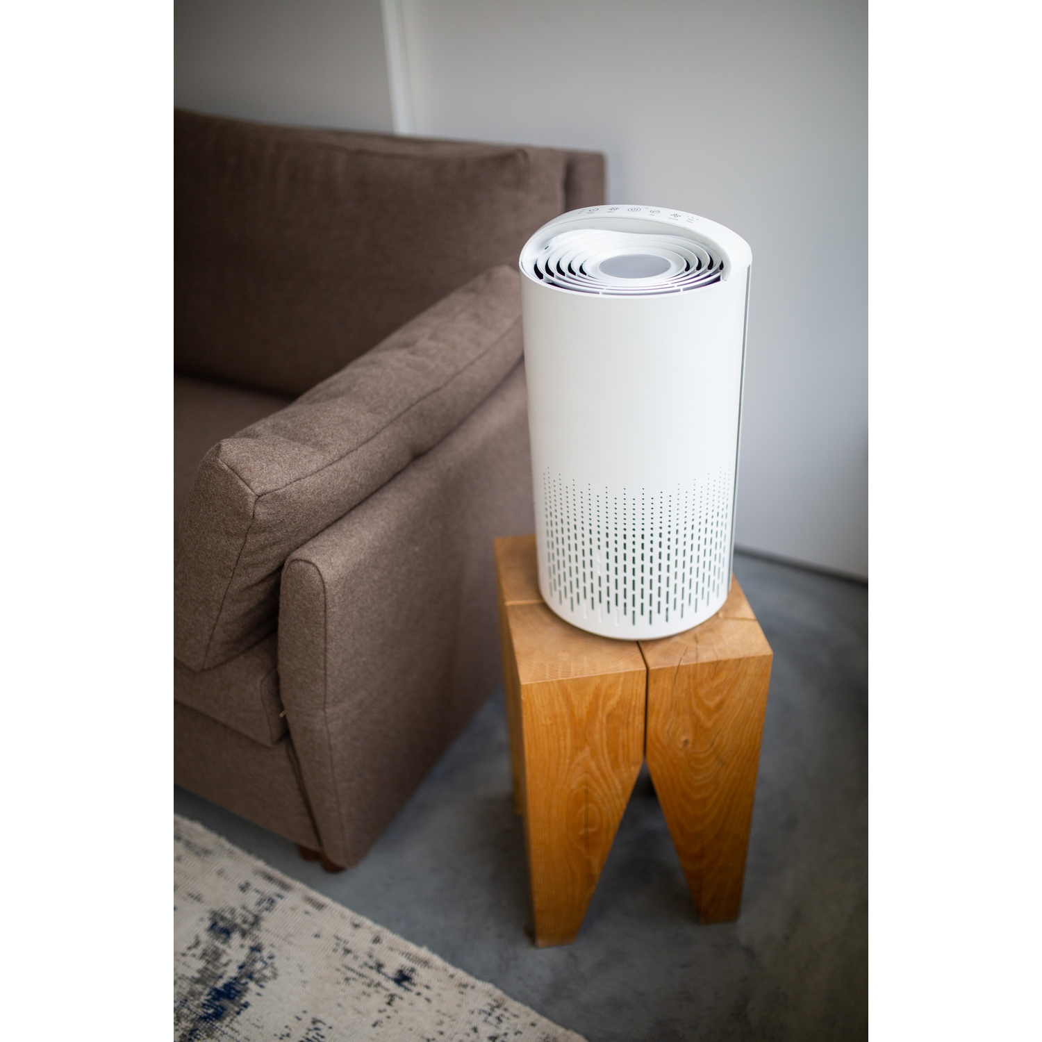 A white air purifier sits on a wooden stool. A brown sofa is to the left. A rug is visible in the foreground.