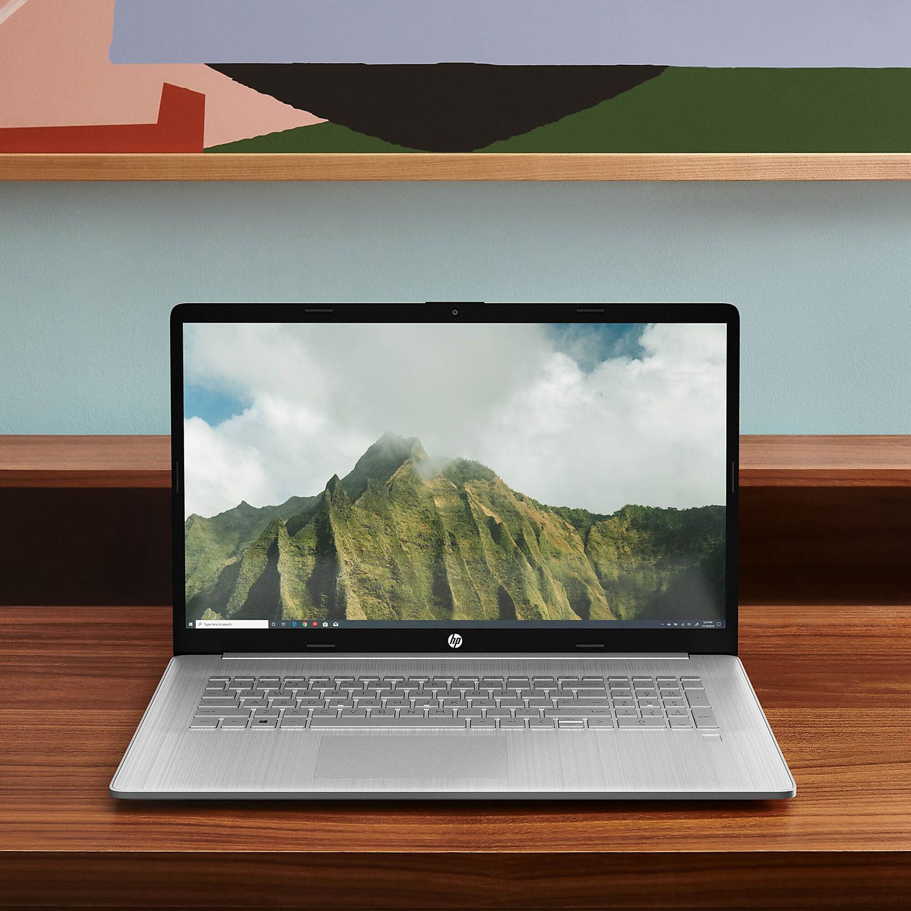 A silver HP laptop displays a mountain scene. It sits on a wood desk, with a colorful wall art in the background.