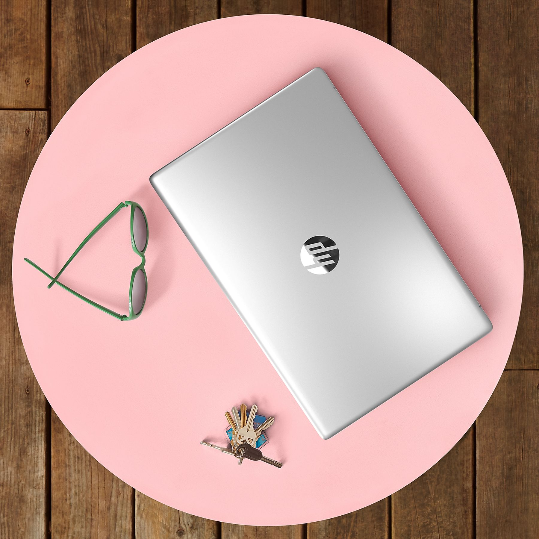 A silver laptop with HP logo, green glasses, and keys on a pink circle, set on a wooden table.