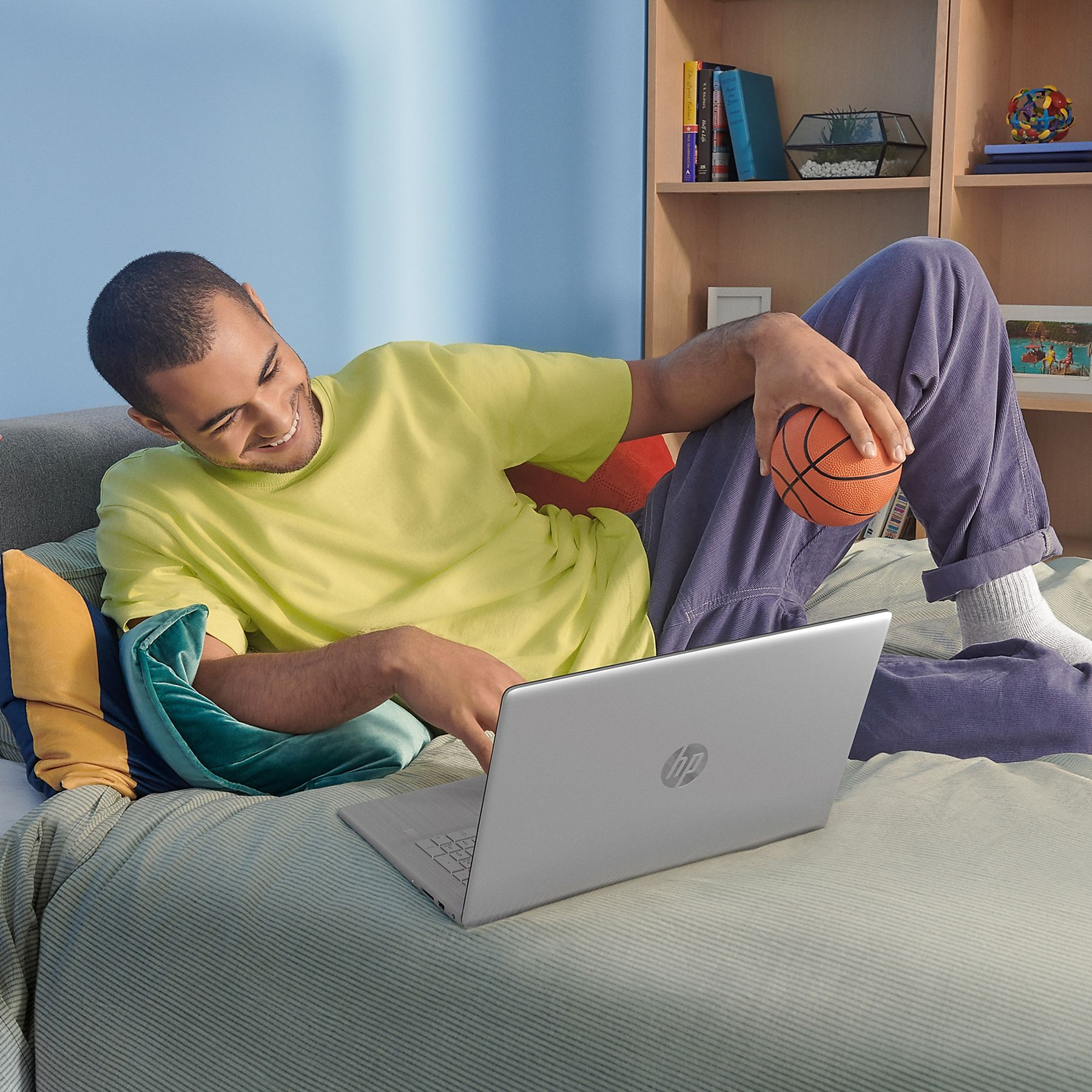 A man lies on a bed with a laptop and a basketball. Wearing a yellow shirt and purple pants, smiling.