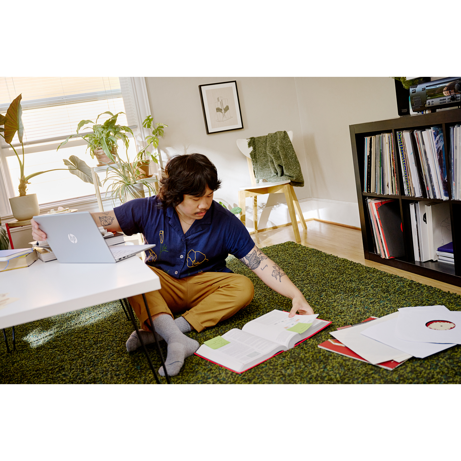 A person sits on a green rug, using a laptop and book. Plants and a record shelf are in the background.