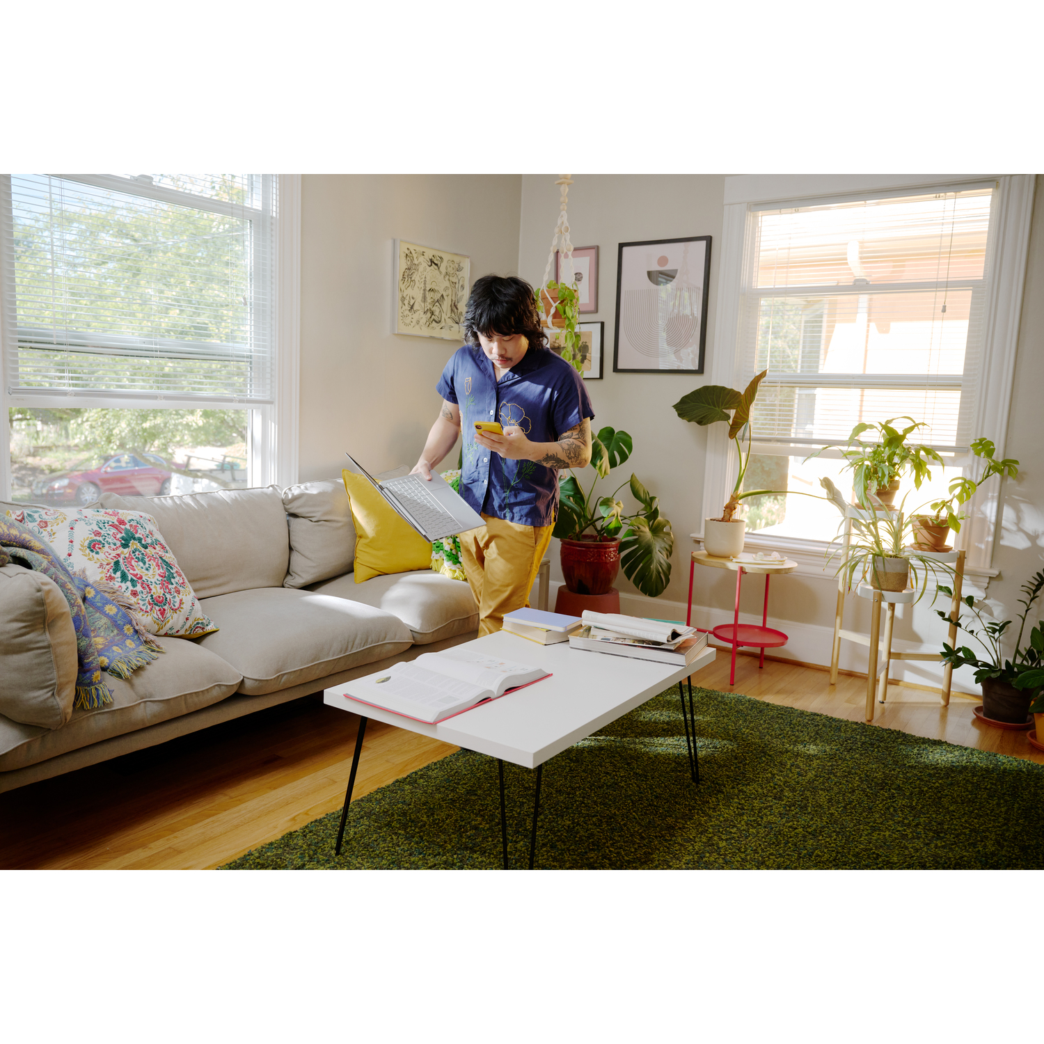 A person in a room with plants and a laptop. Holding a phone, the person stands near a white table with an open book.