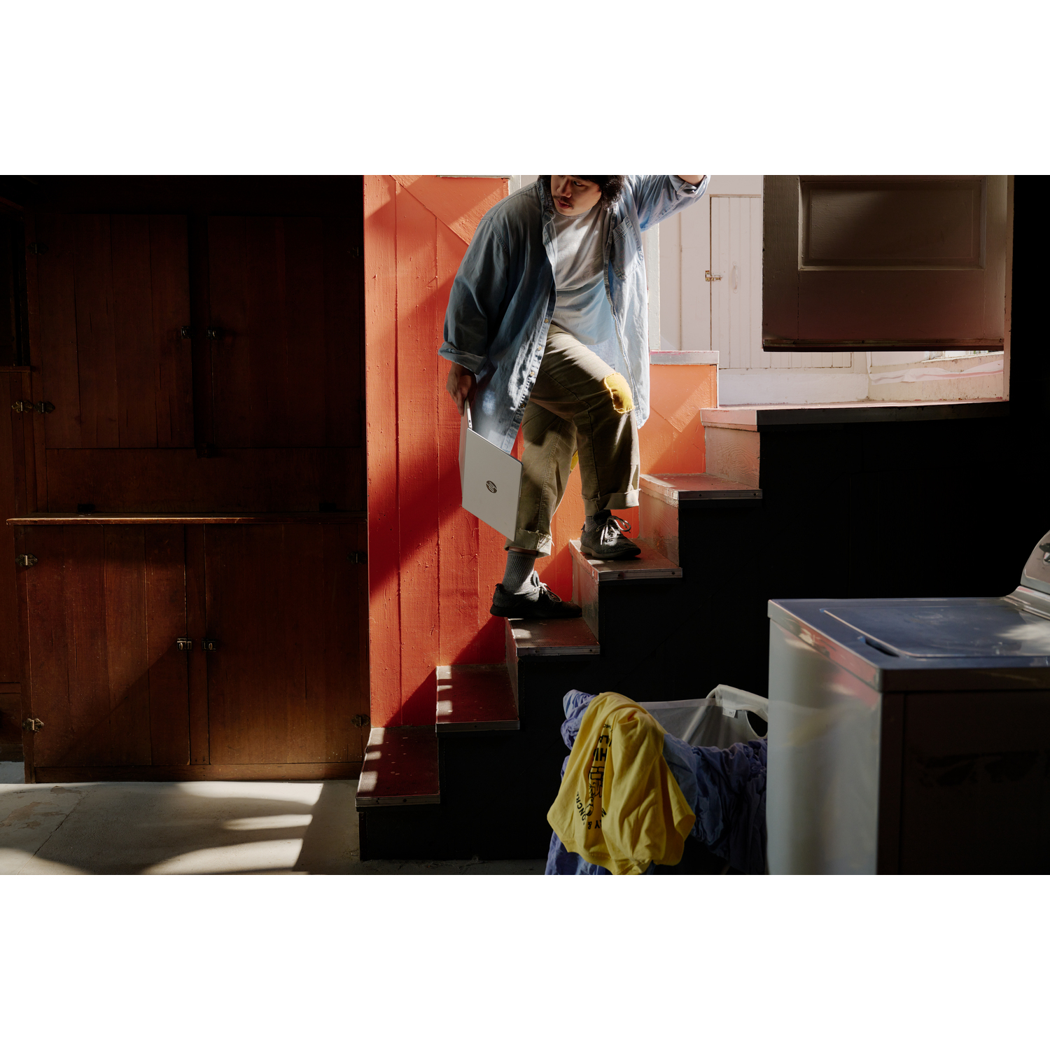 A person descends stairs holding a laptop. Laundry and a washing machine are visible. Wooden cabinets and a red wall are in the background.