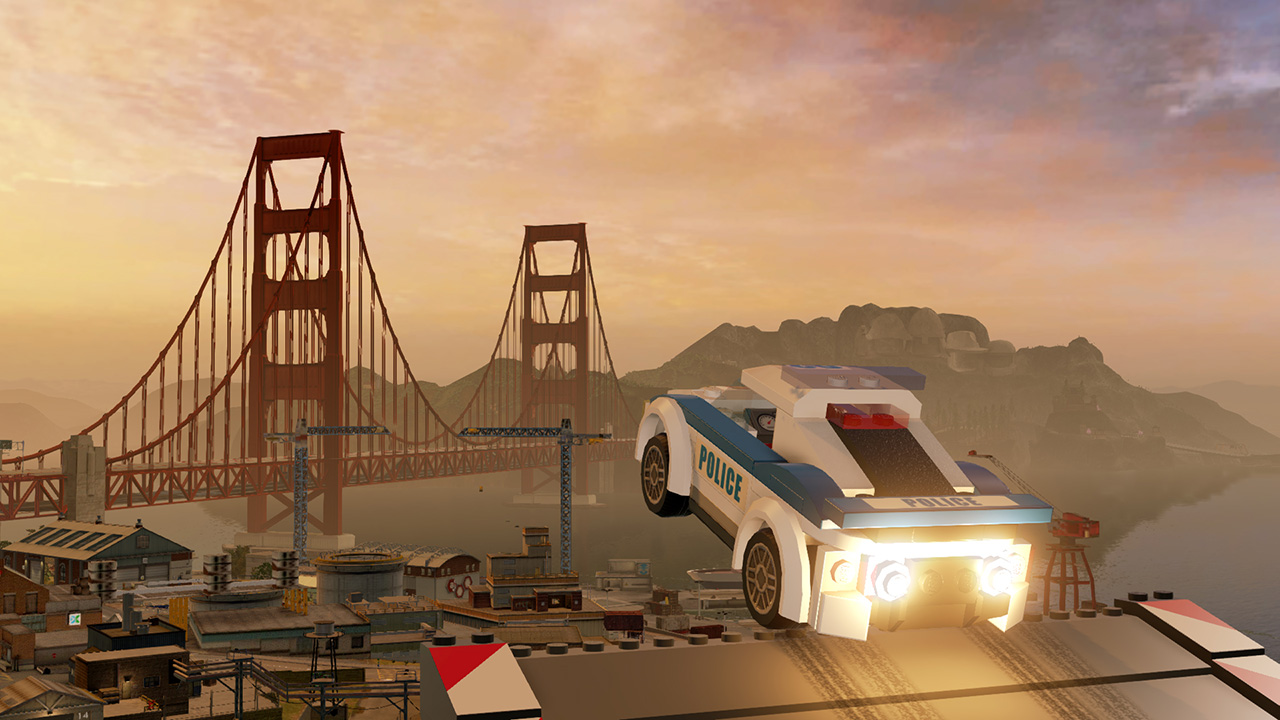 Lego-Polizeiauto springt in der Nähe der Golden Gate Bridge. Gebäude und Wasser im Vordergrund, Berge und Sonnenuntergang im Hintergrund.