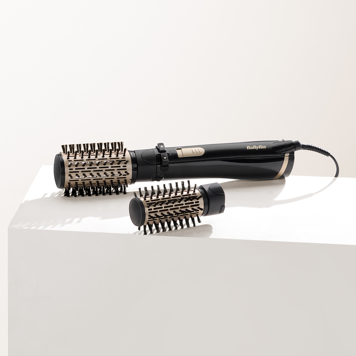 A black and gold Babyliss hair styler with two brush attachments on a white surface. The brand name is visible.