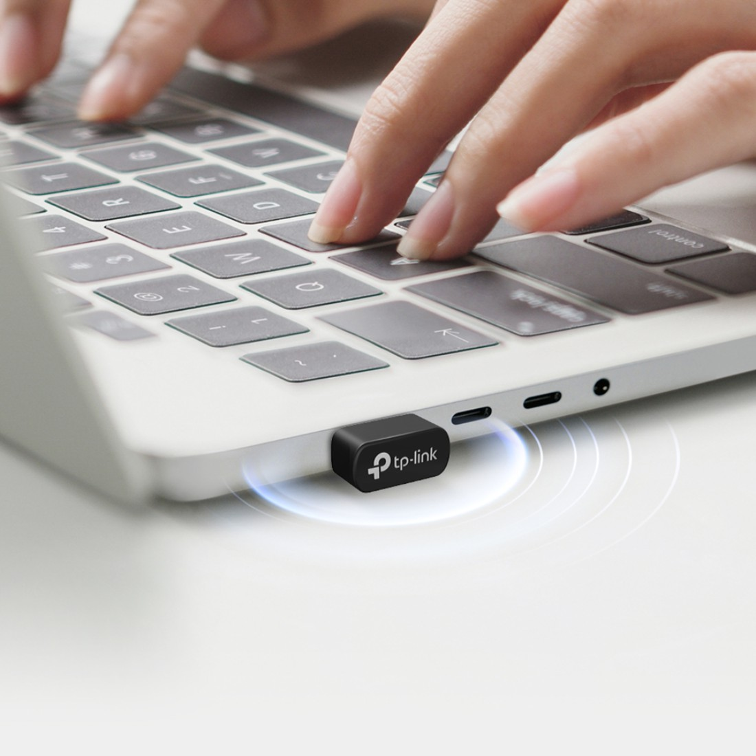 Hände tippen auf einer Laptop-Tastatur. Ein schwarzer TP-Link USB-Adapter ist eingesteckt. Weiße Oberfläche mit Tastatur und Händen.