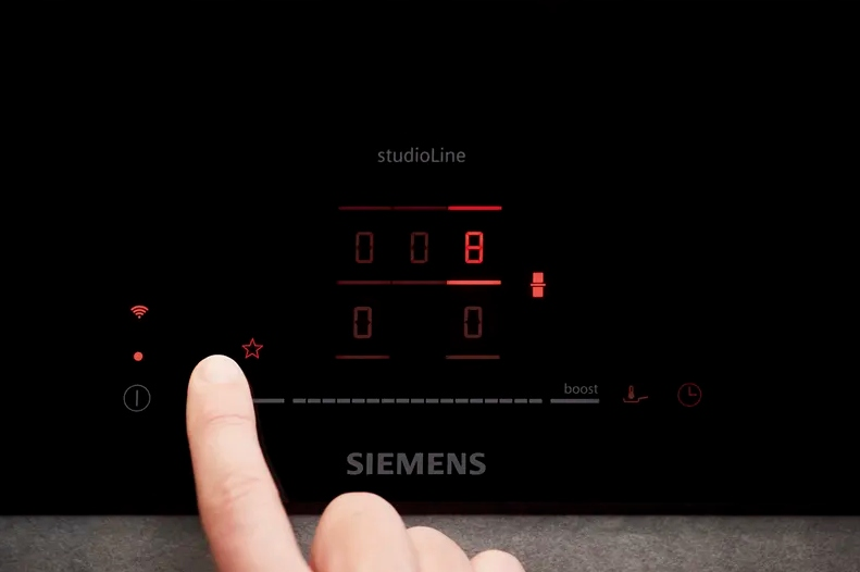 Palec naciska przycisk w kształcie gwiazdy na czarnej płycie grzewczej Siemens. Czerwone cyfry pokazują 008. Widoczne słowo 'studioLine'.