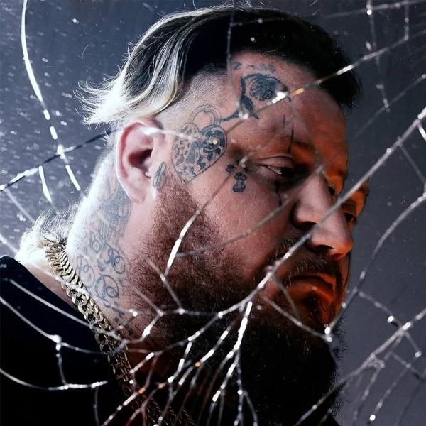 Mann mit Tätowierungen und Goldkette hinter zerbrochenem Glas. Er hat einen Bart und kurzes, helles Haar. Dunkler Hintergrund.