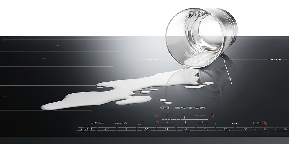 Szklanka mleka wylewa się na czarną płytę indukcyjną Bosch. Mleko rozlewa się po powierzchni.