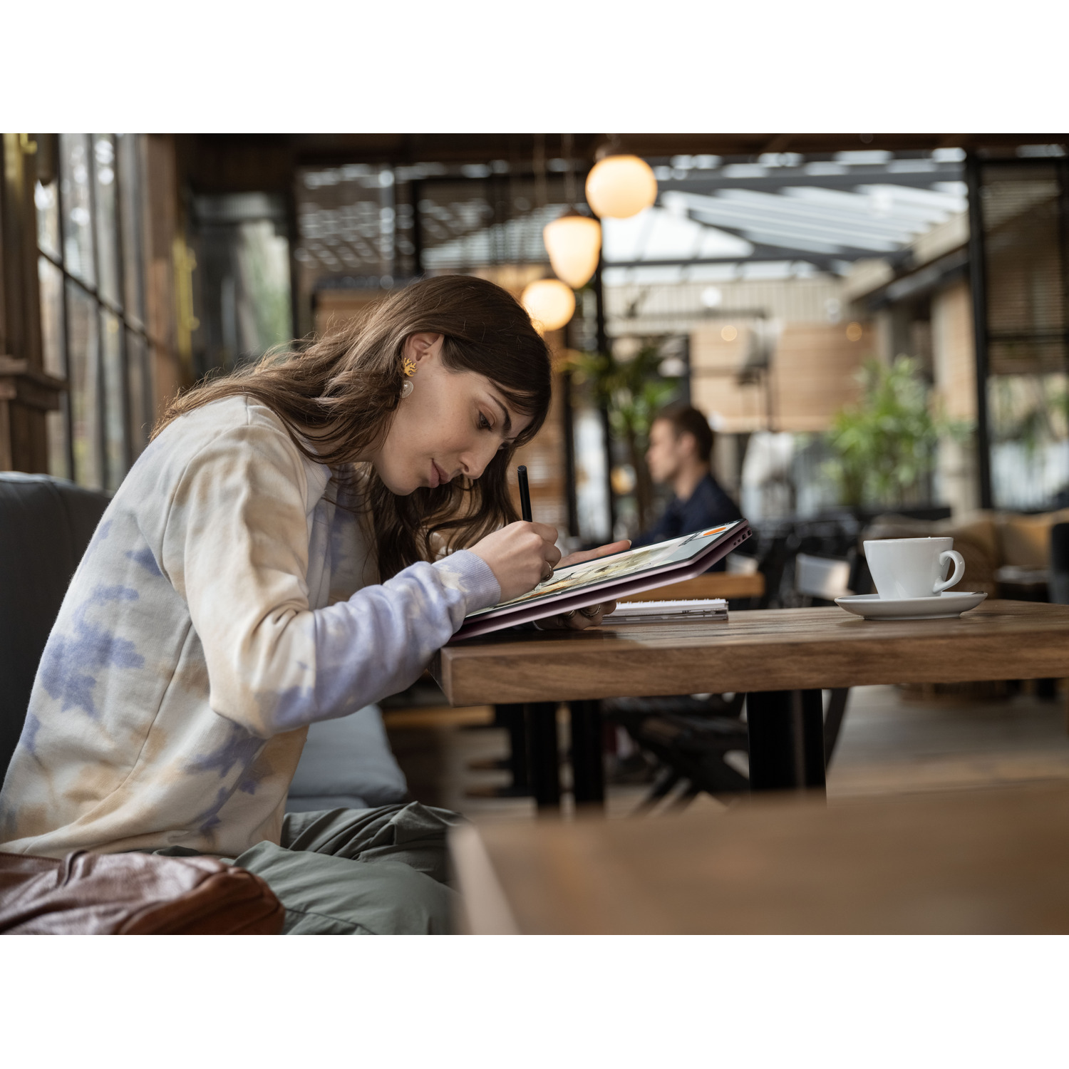 Woman in cafe drawing on a tablet. Cup and saucer on table. Another person in background. Lights hanging from ceiling.