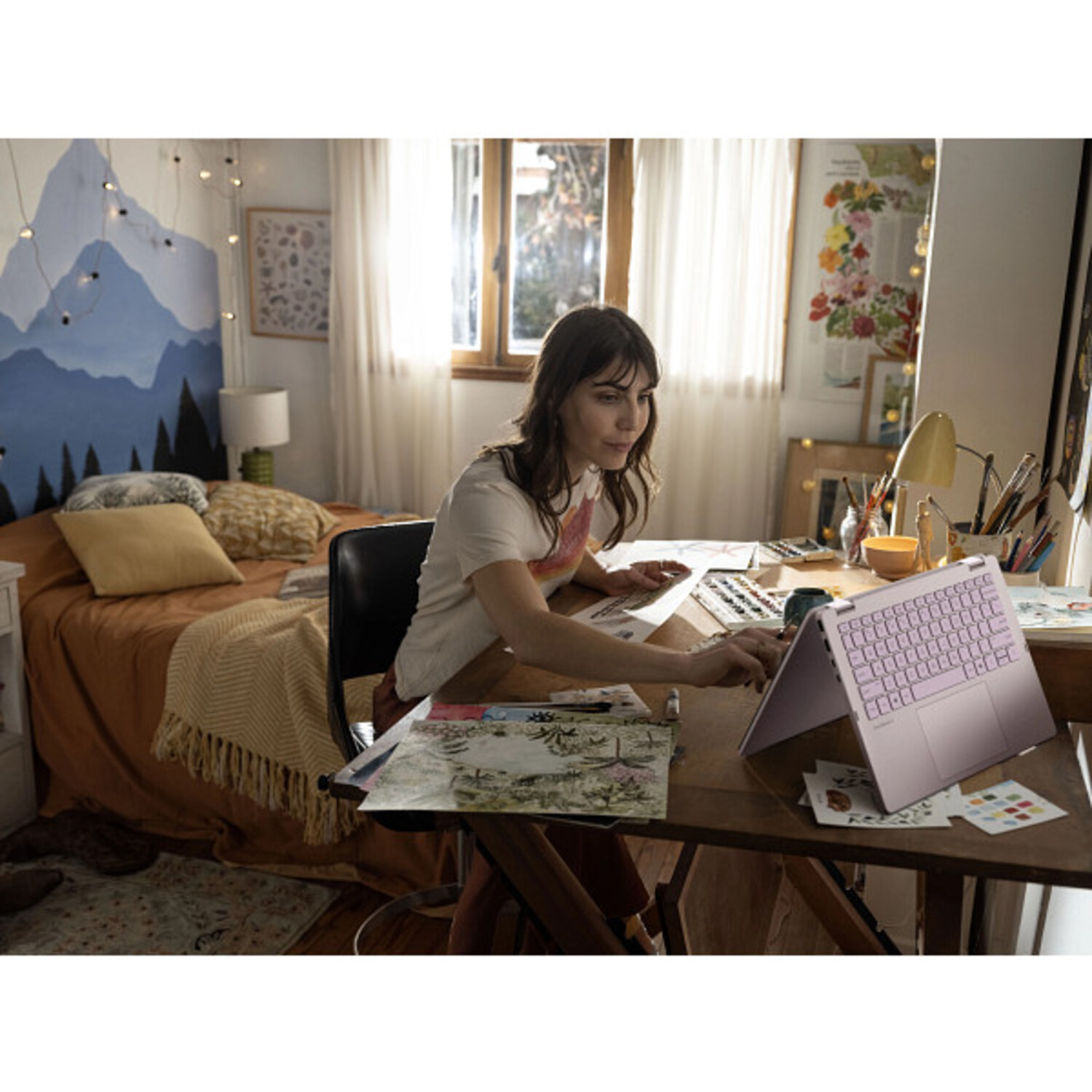 Woman working on a laptop at a desk in a room with a bed and window. There are art supplies and drawings on the desk.
