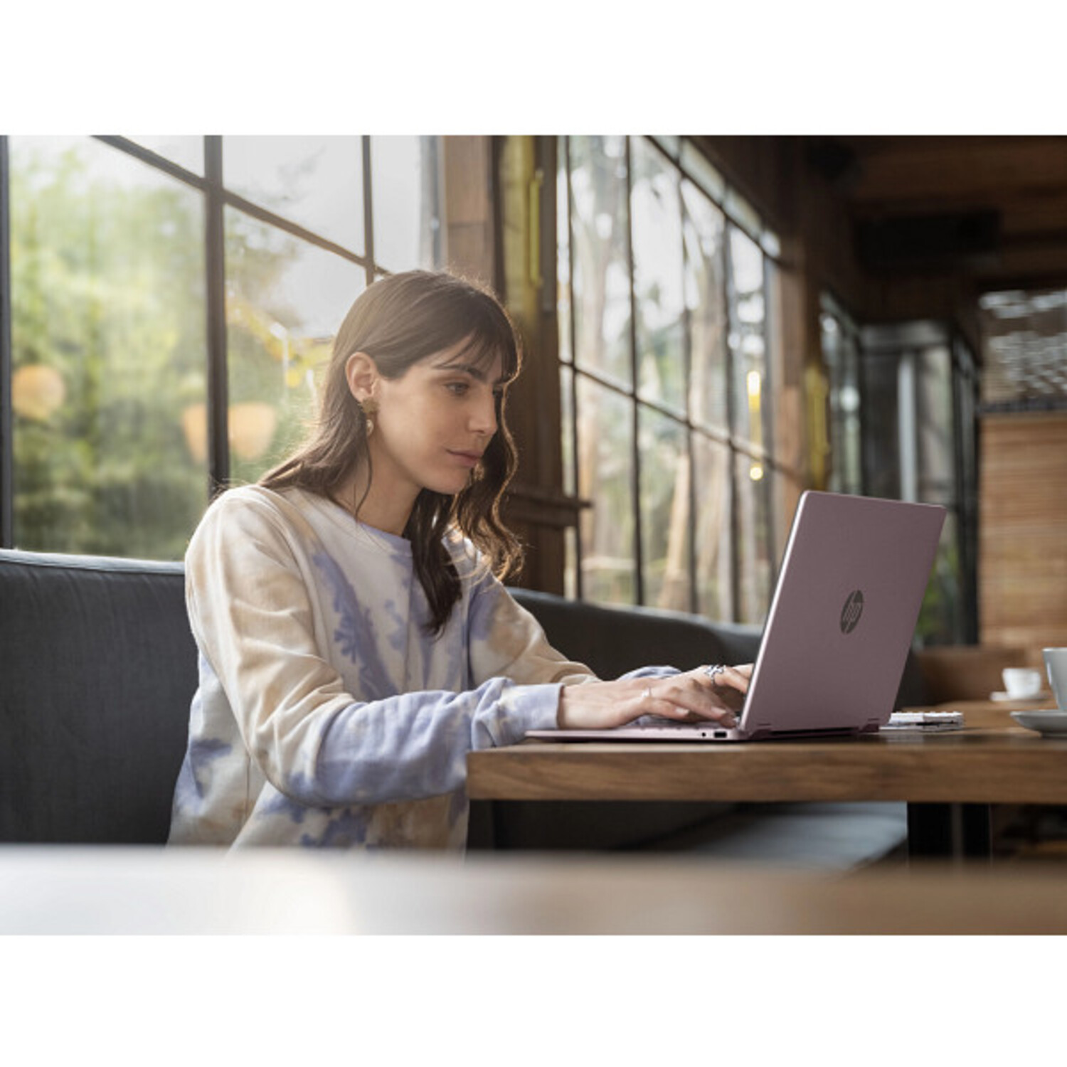 A woman works on a pink HP laptop at a wooden table. She wears a tie-dye sweater. The background shows a window.