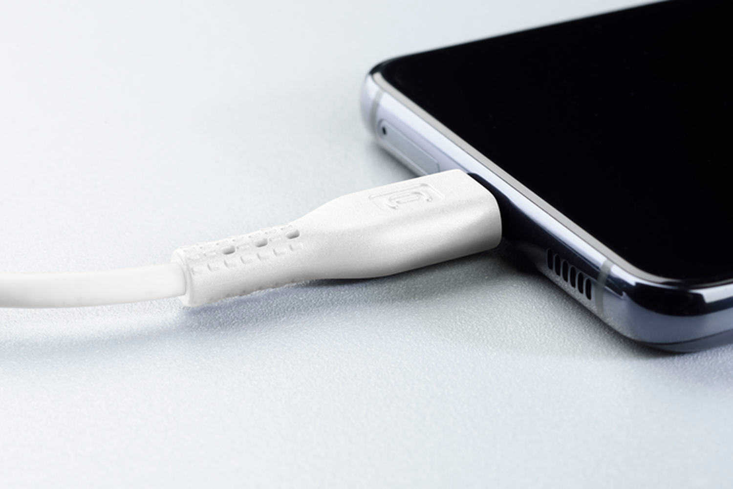 Un cable de carga blanco está conectado a un teléfono inteligente negro. El cable está sobre una superficie gris claro.