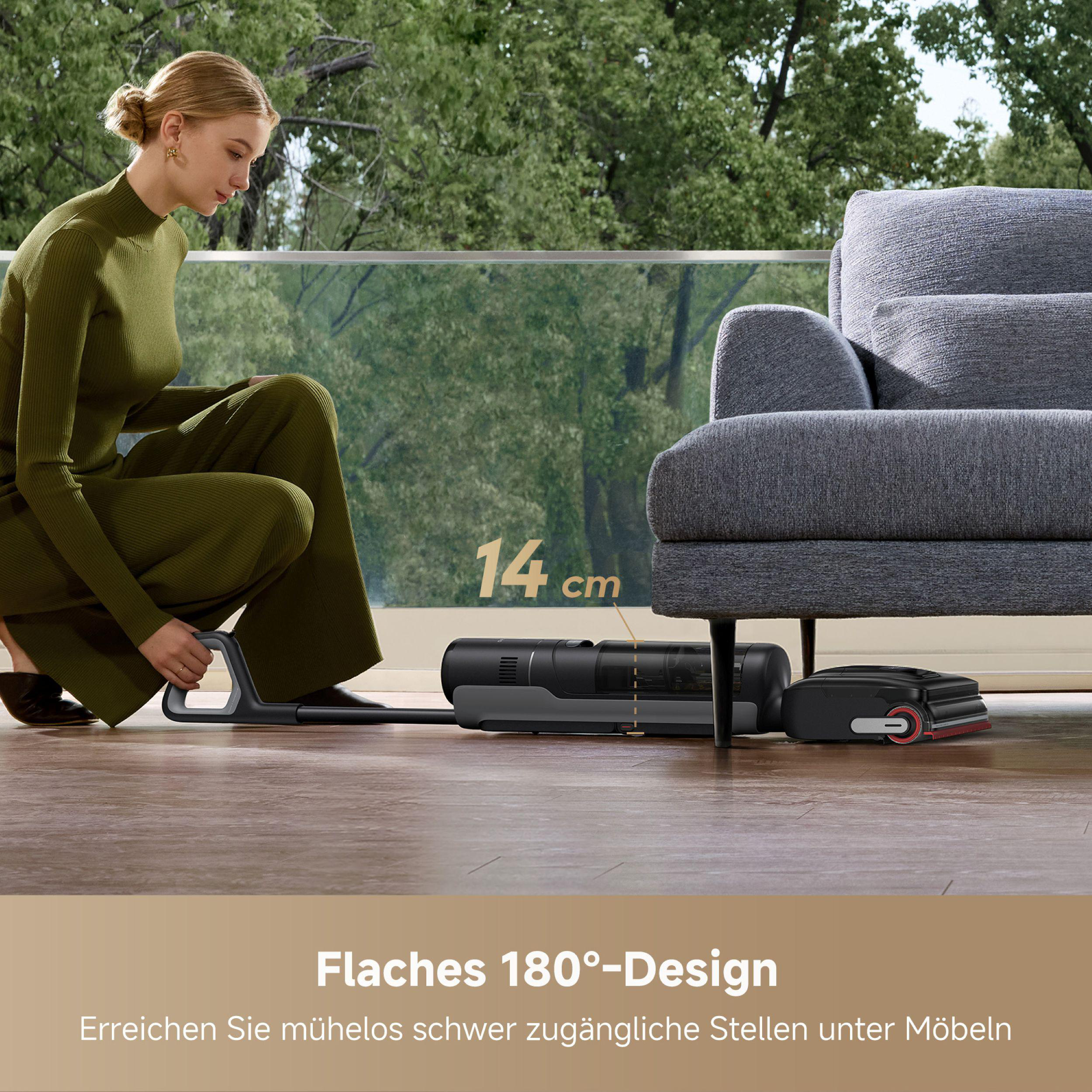 Eine Frau in Grün benutzt einen Staubsauger. Im Hintergrund sind ein Sofa und Bäume. Der Reiniger ist grau und schwarz.