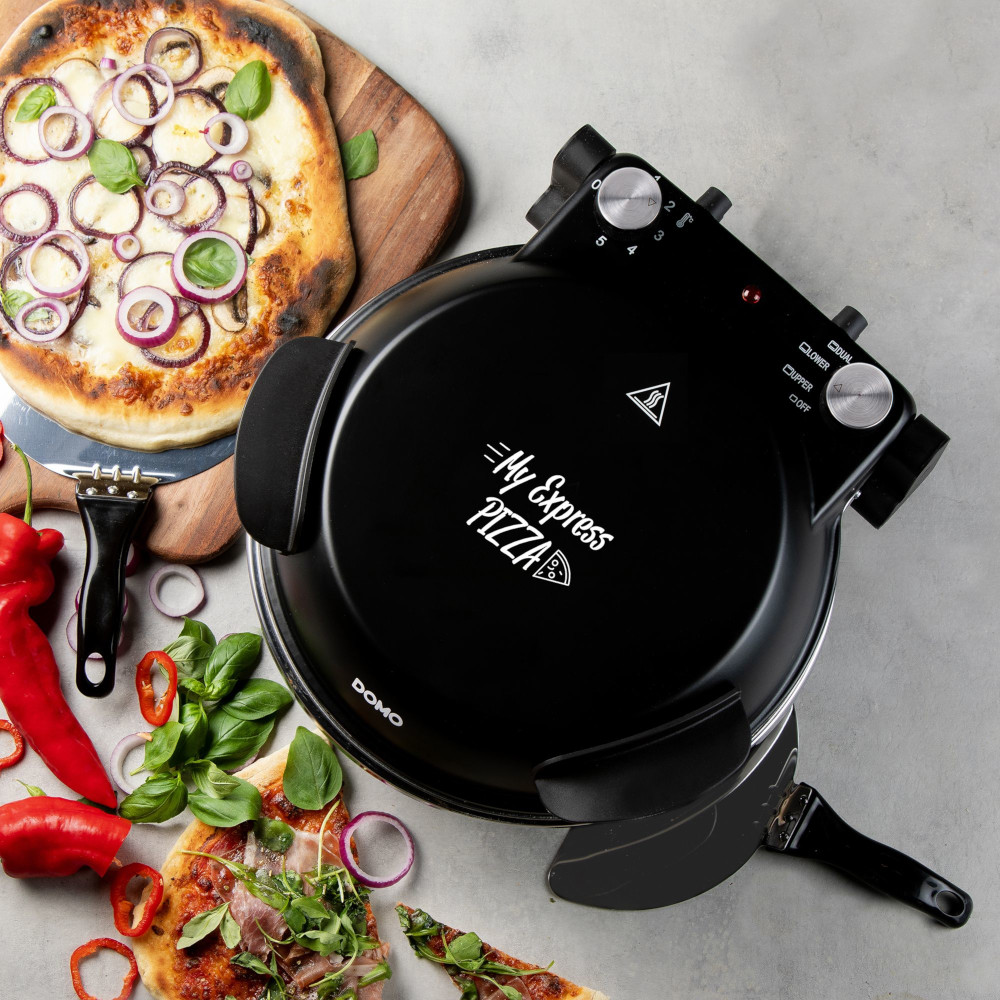 A black pizza oven, pizzas with toppings, and ingredients are on a gray surface. Red pepper slices and basil leaves are scattered.