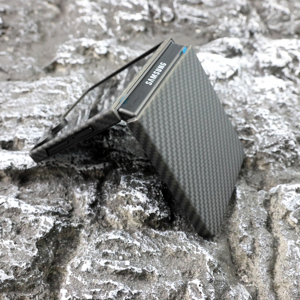 Egy félig nyitott Samsung összecsukható telefon szürke, texturált sziklafelületen. A telefon karbonminta borítású tokkal rendelkezik.