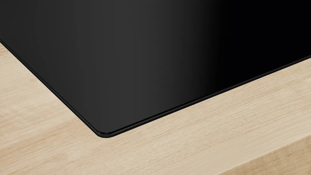 Zbliżenie czarnej, prostokątnej płyty grzejnej na jasnej drewnianej powierzchni. Krawędź płyty jest lekko zakrzywiona.