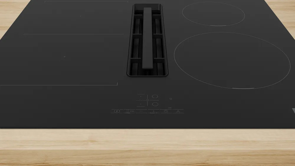 Czarna płyta grzewcza ze zintegrowanym wentylatorem. Widoczne są dwie okrągłe strefy gotowania. Płyta grzewcza spoczywa na jasnej drewnianej powierzchni.