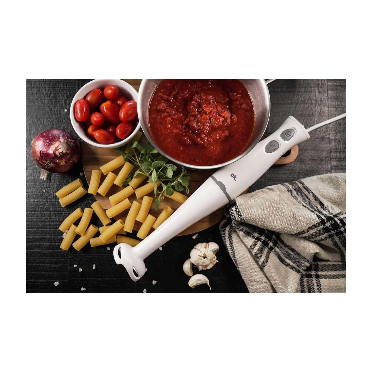 Ingredienti per la pasta: pomodori, pasta, salsa, cipolla, aglio, erbe aromatiche e un frullatore. Tutto su un tagliere e una superficie nera.