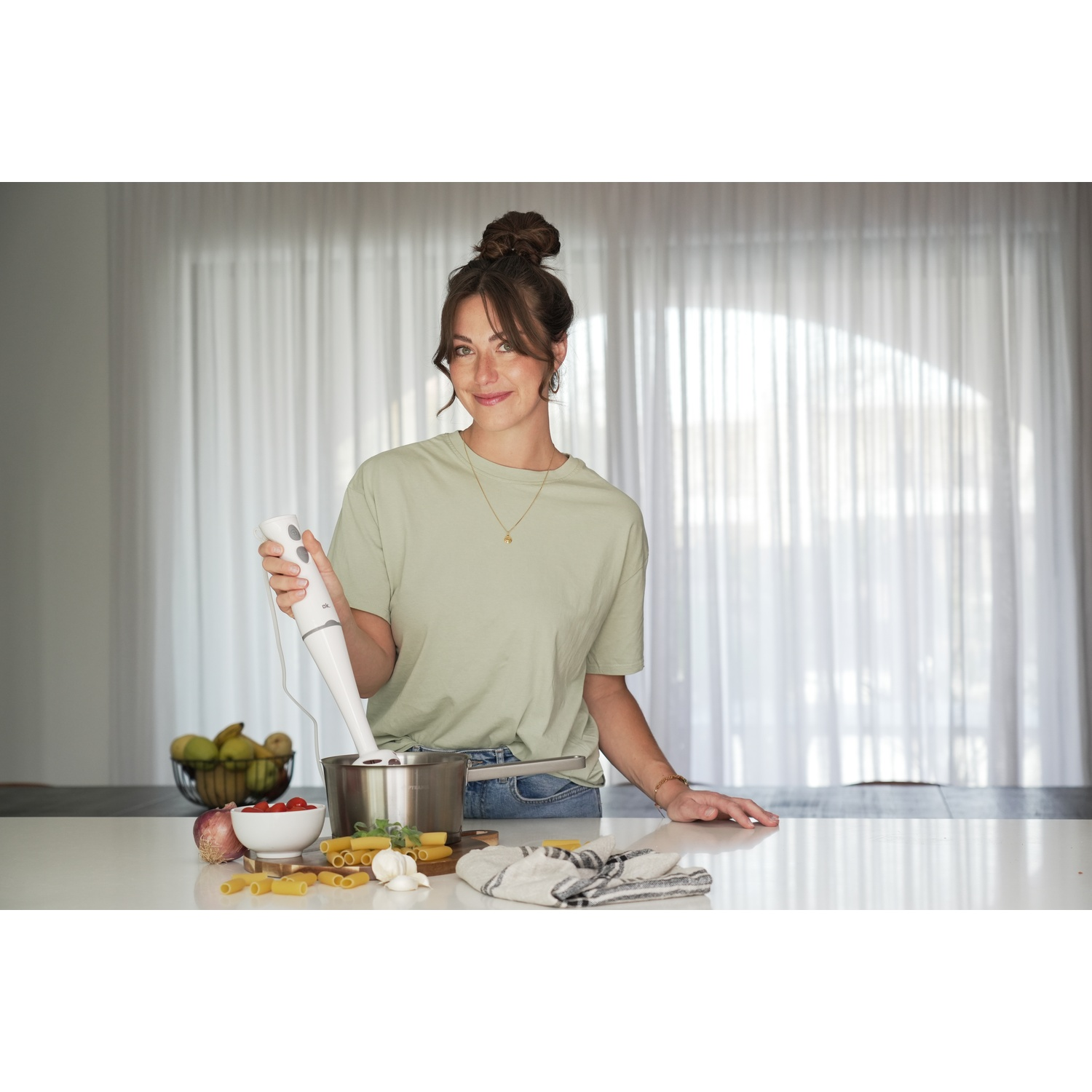 Donna in cucina con un frullatore a immersione. Ingredienti sul bancone: pomodori, pasta, aglio e una ciotola di frutta.