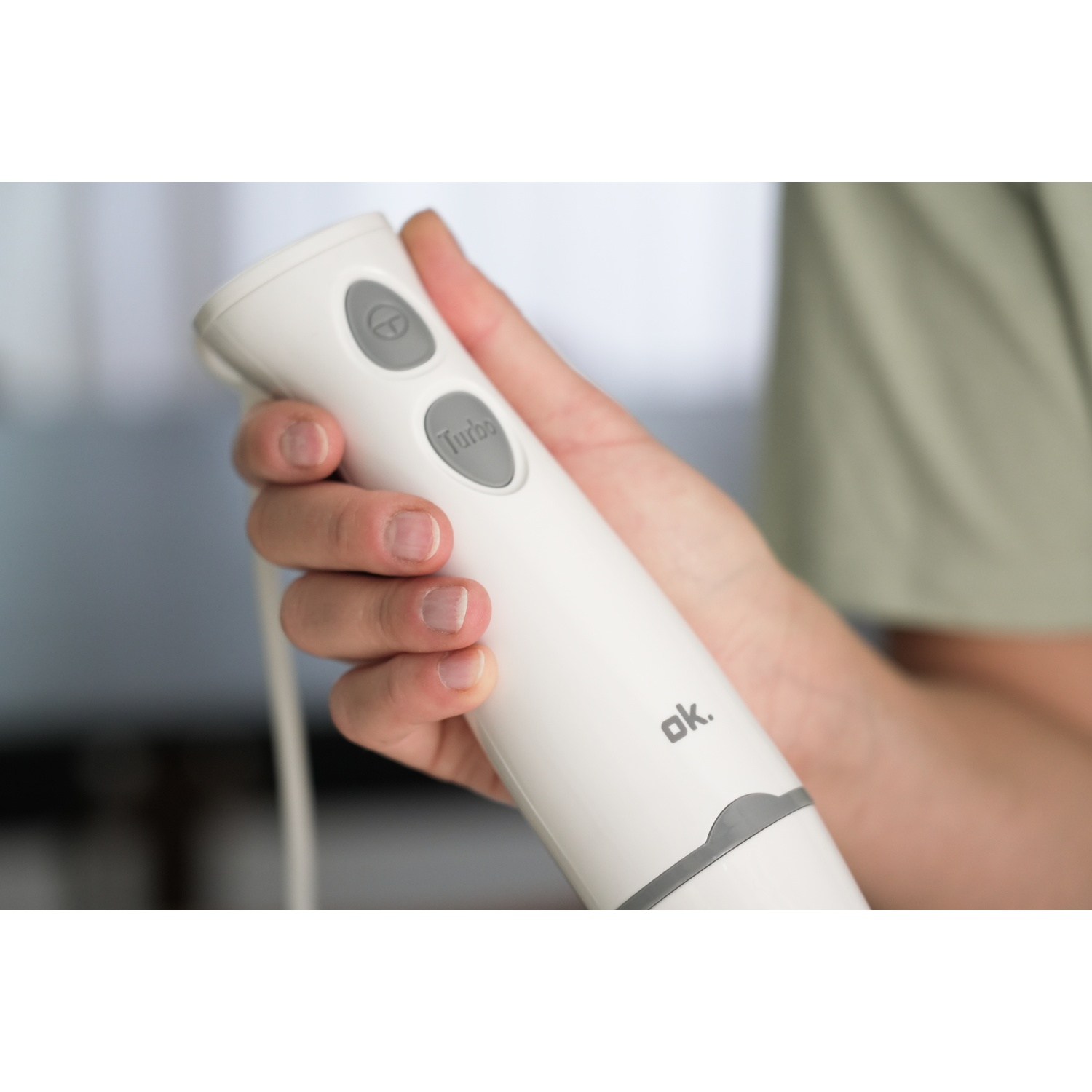 Una mano tiene un frullatore a immersione bianco con pulsanti grigi. La parola "Turbo" è visibile. Una persona indossa una camicia verde.